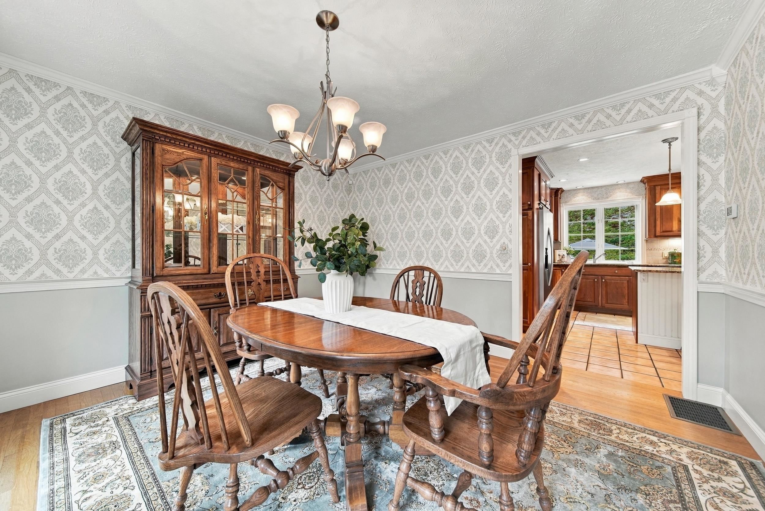 Dining room with an oval wooden table, glass-front china cabinet, patterned wallpaper, and chandelier.