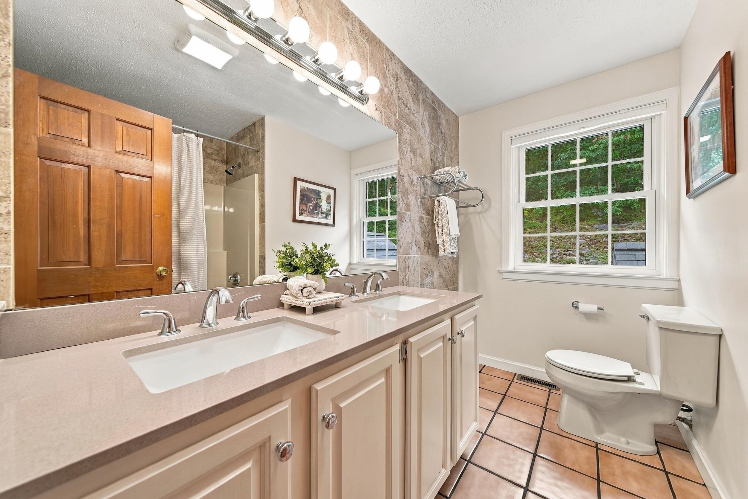 Bathroom with a double-sink vanity, large mirror, tiled shower, and window overlooking woods.