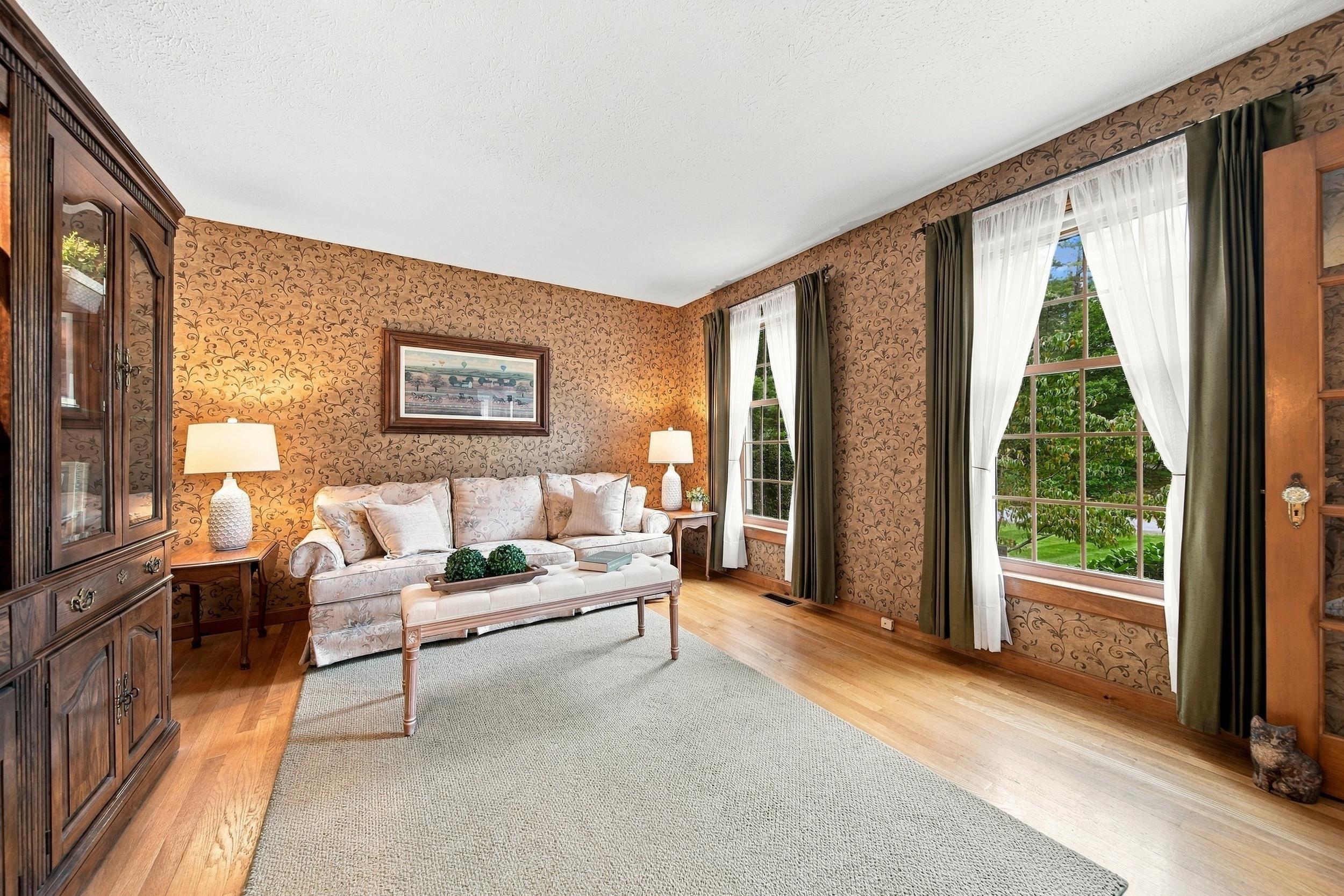 Living room with patterned wallpaper, pale sofa, hardwood floors, and tall curtained windows overlooking greenery.
