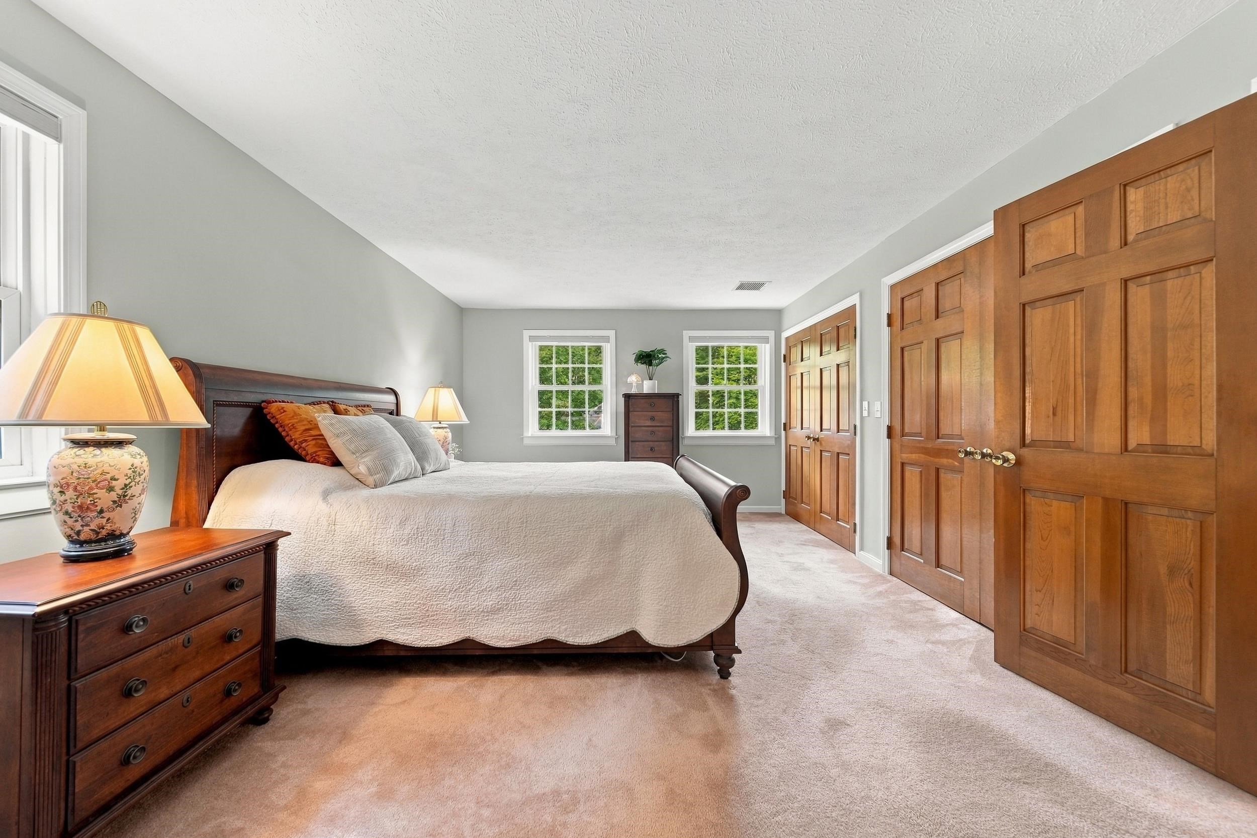 Spacious bedroom with cream bedding, dark wood furniture, two windows, and paneled wooden closet doors.