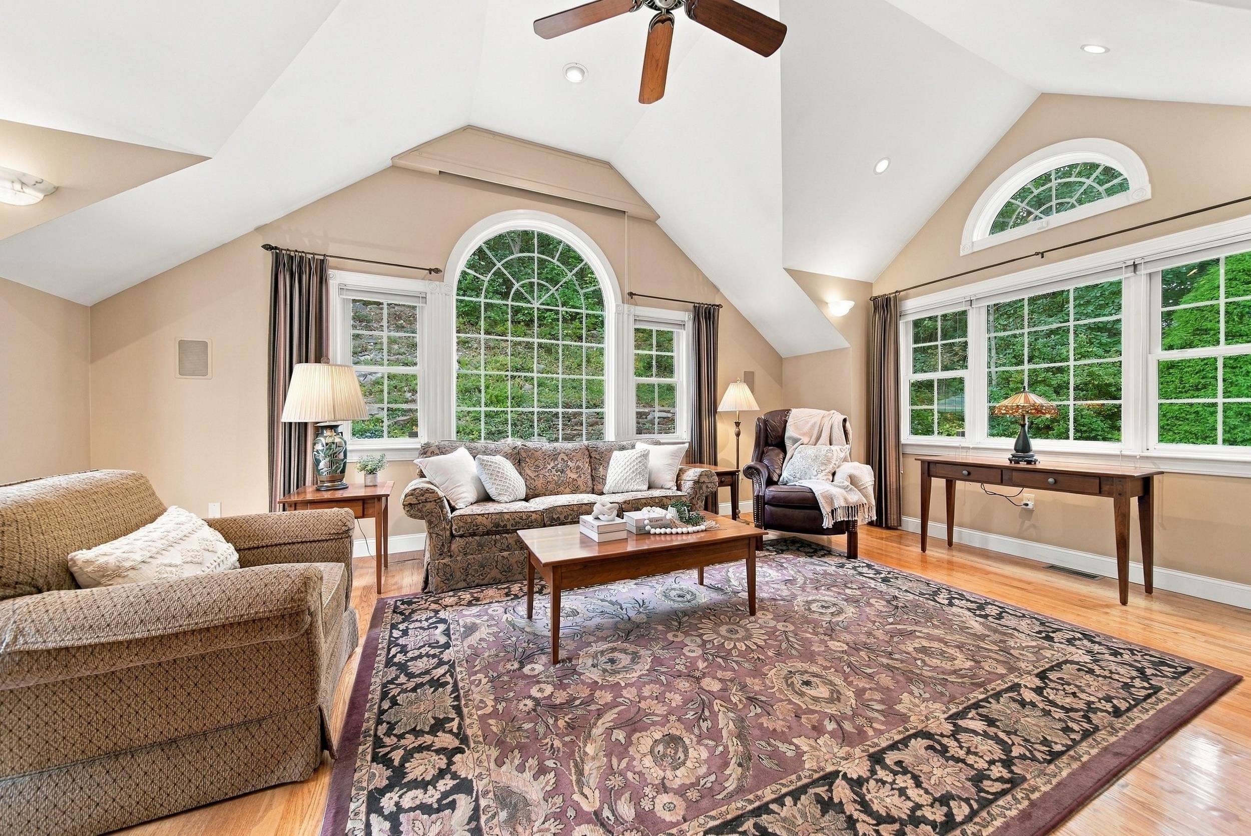 Furnished living room with vaulted ceiling, arched windows, wooded views, and a large patterned rug.