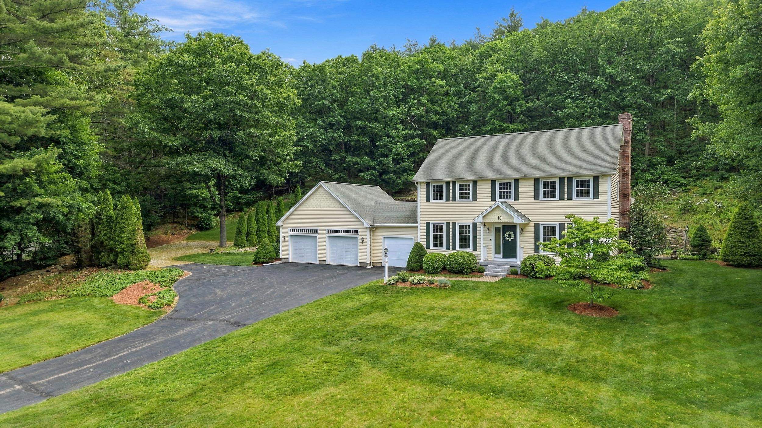 Cream two-story home with three-car garage, landscaped lawn, and woods behind it.