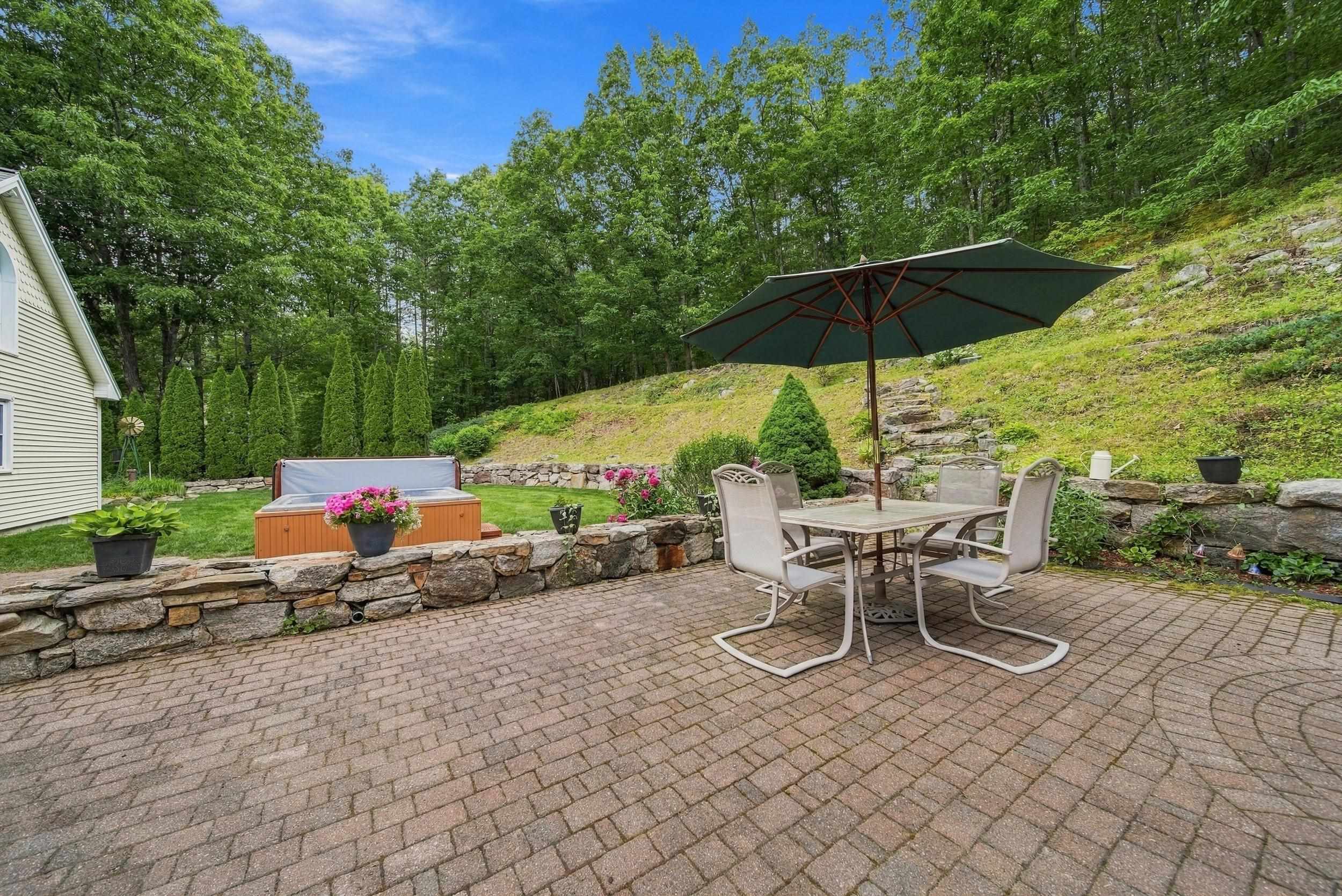 Brick-paver patio with umbrella-covered dining set, hot tub, and stone walls beside a wooded hillside.