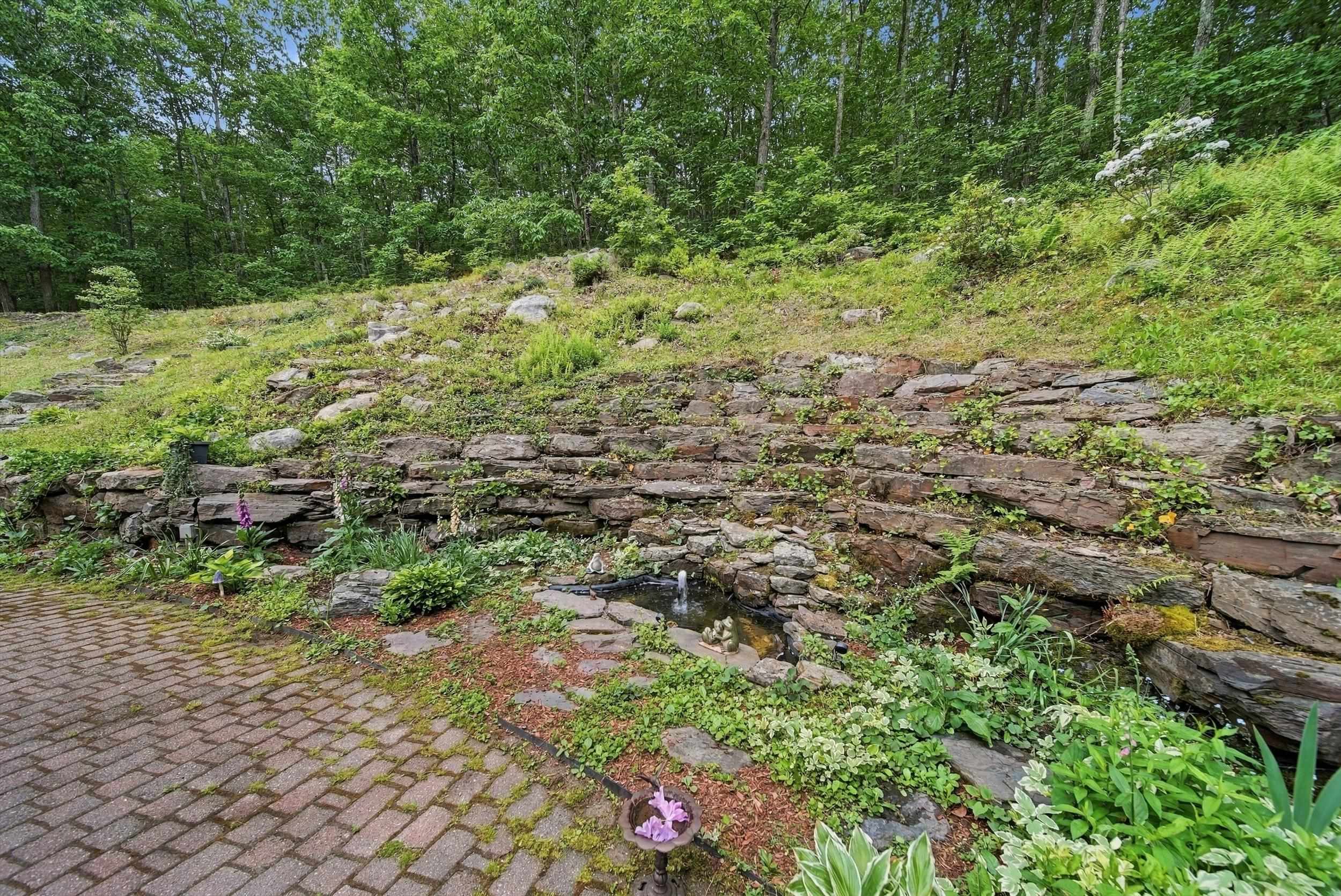 Landscaped yard with a stone retaining wall, small fountain pond, flowering plants, and brick patio.