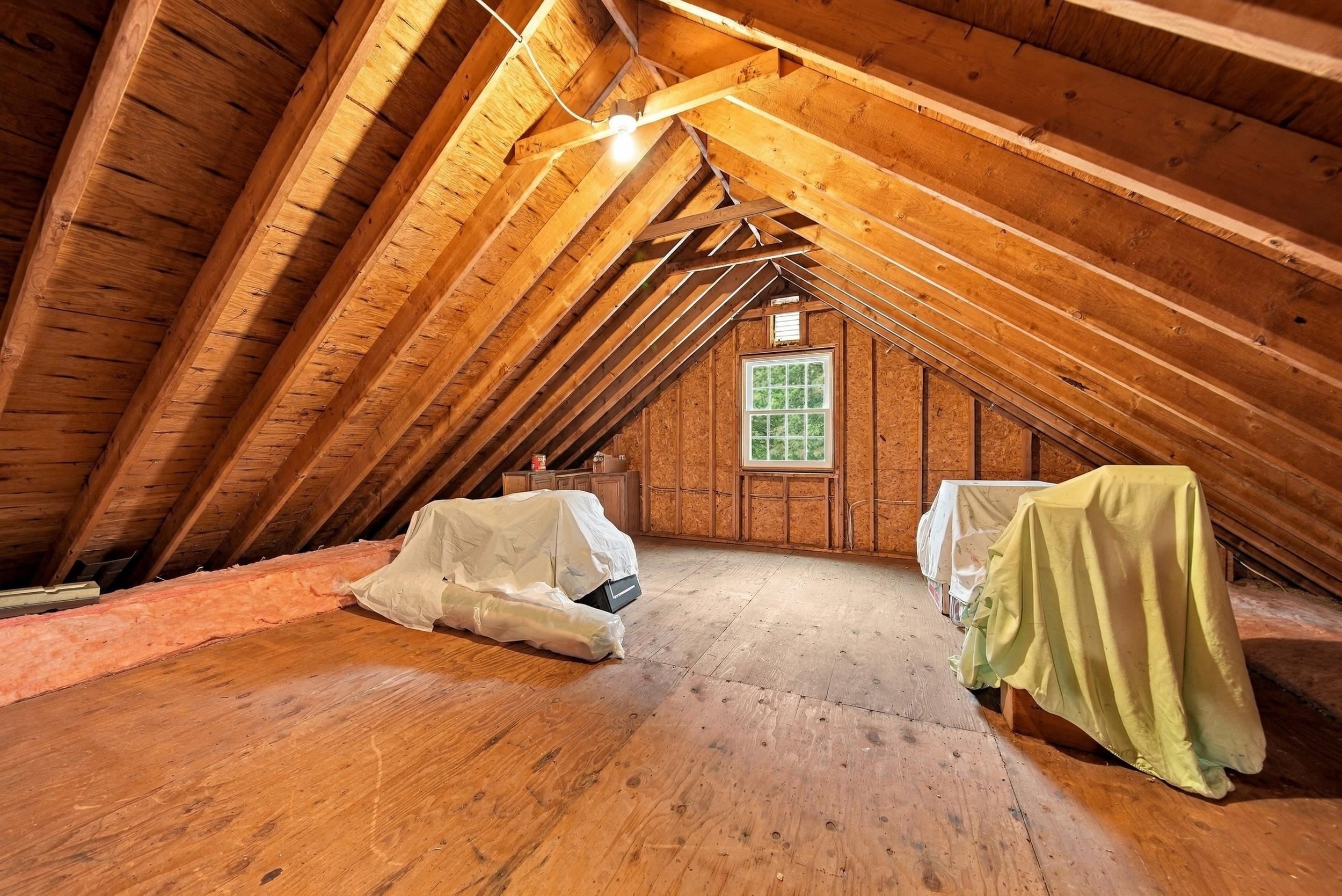 Unfinished attic with exposed wooden rafters, insulation along the eaves, and covered items beneath a centered window.