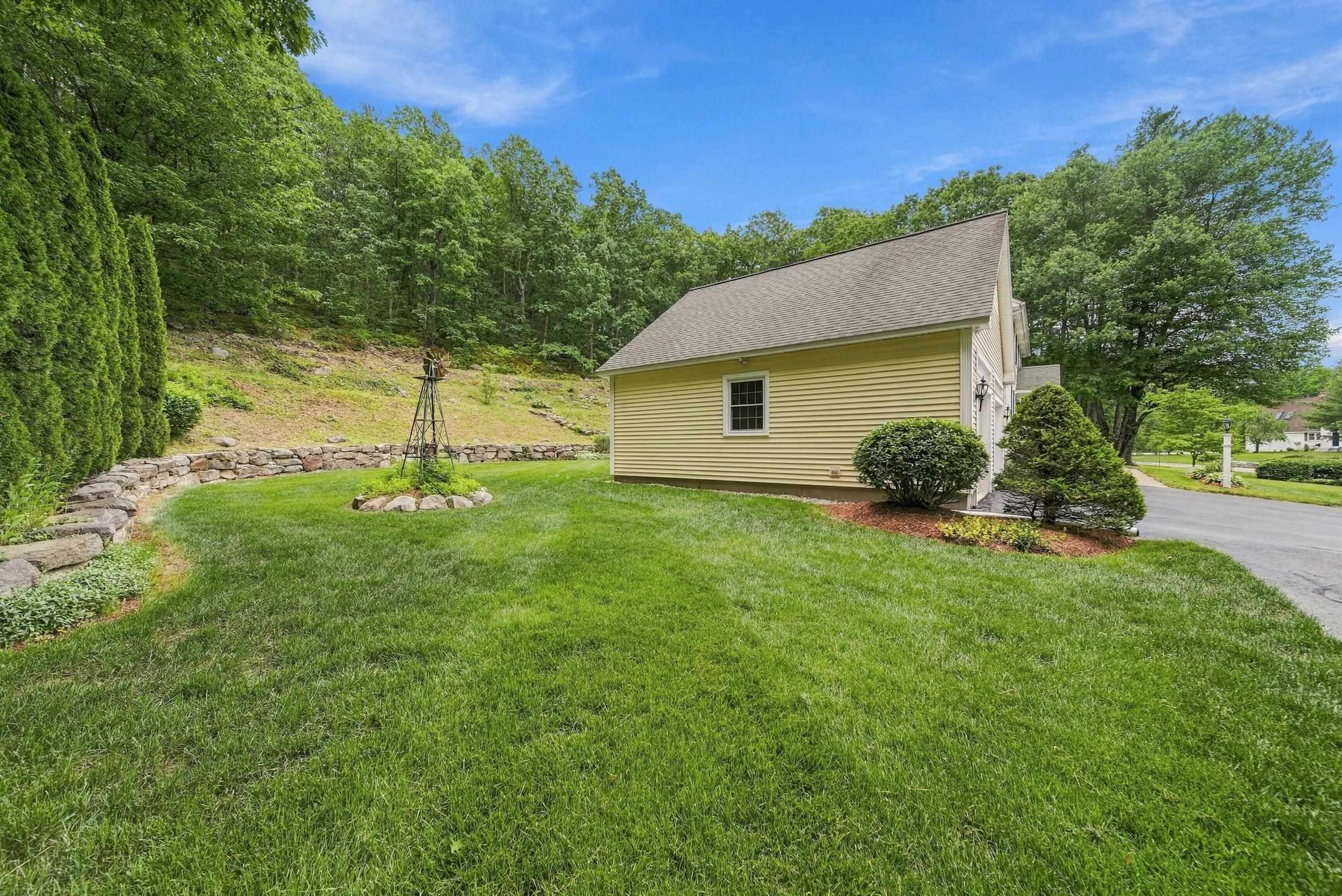 Yellow-sided home beside a landscaped lawn, stone retaining wall, and wooded hillside.
