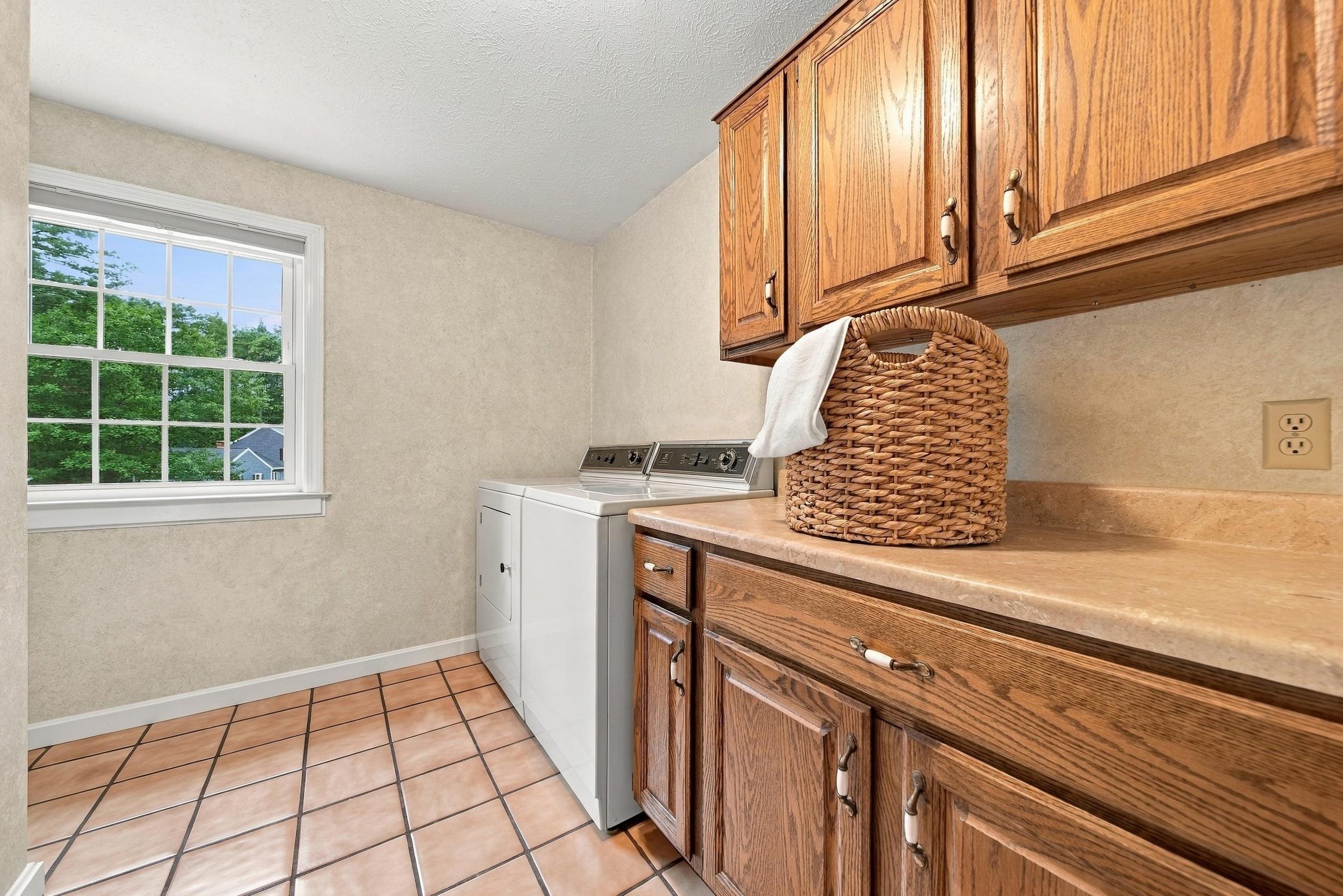 Laundry room with white washer and dryer, oak cabinetry, terracotta tile, and a window overlooking trees.