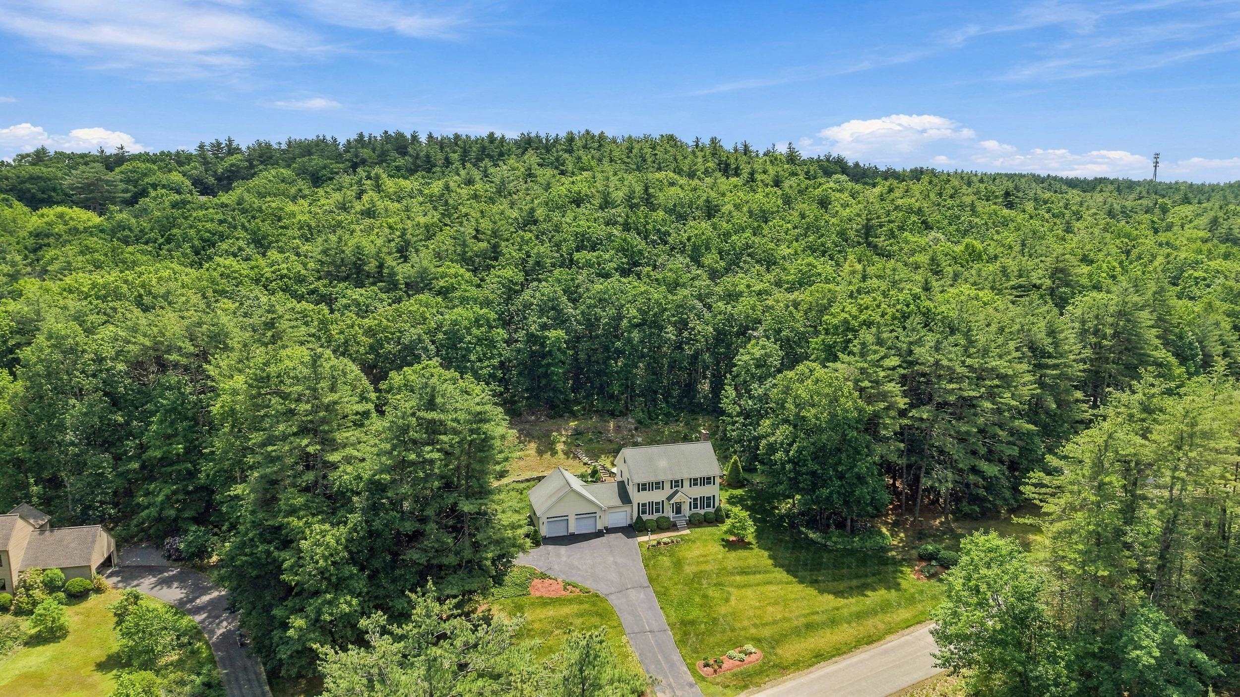Aerial view of a two-story home with an attached garage, lawn, and surrounding woods.
