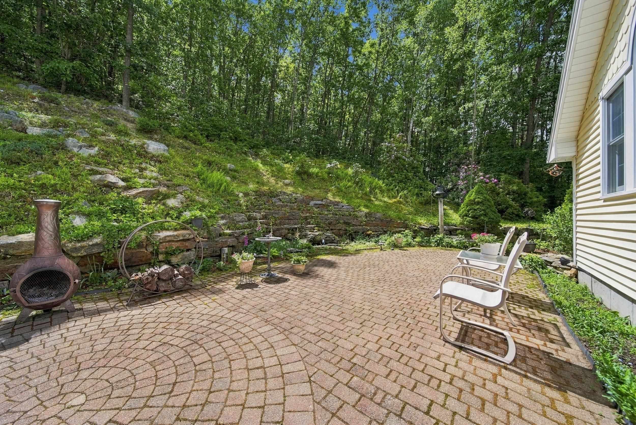 Brick-paver patio with white chairs and a chiminea beside a wooded hillside.