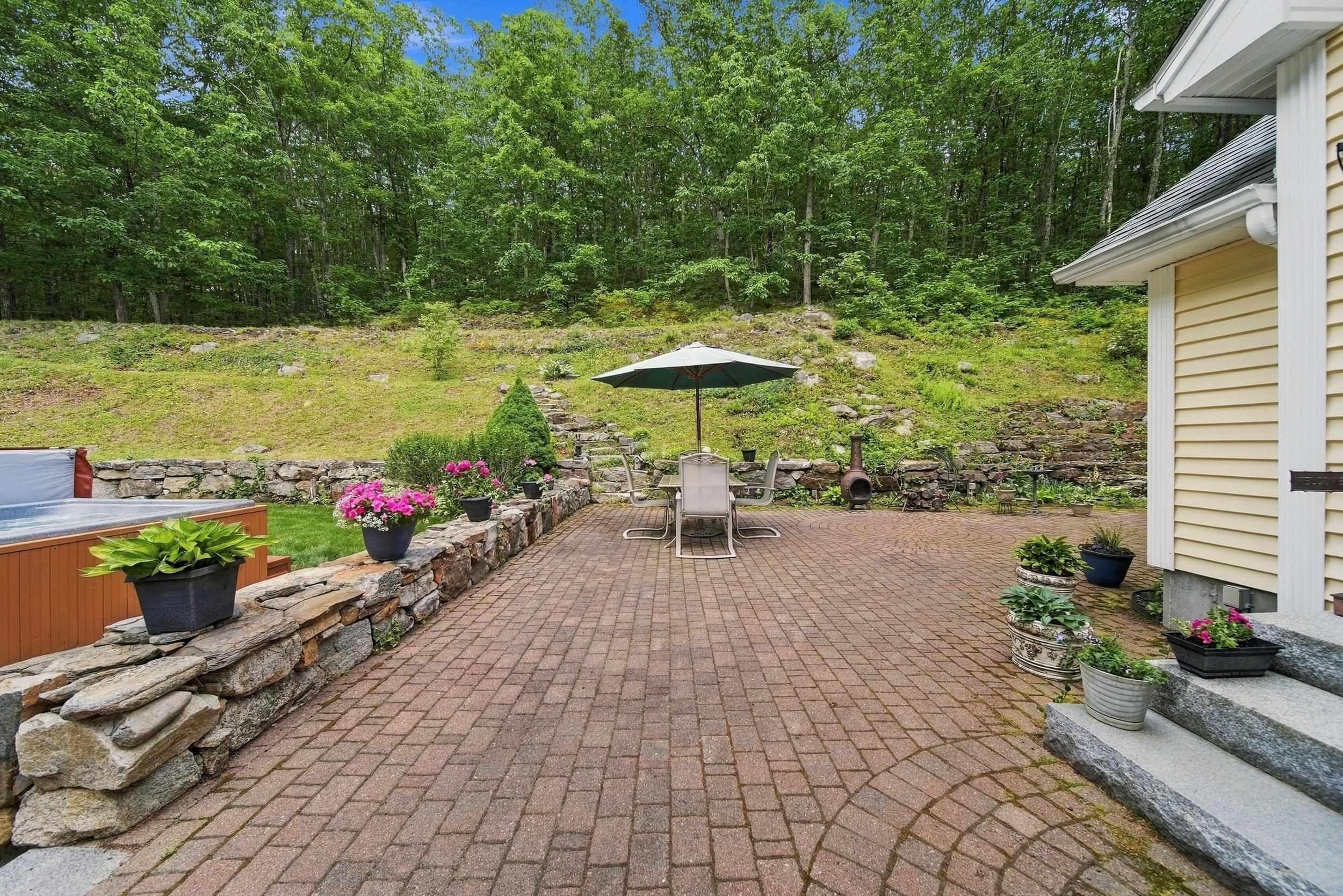 Brick patio with umbrella-shaded dining set, hot tub, stone wall, and wooded hillside.