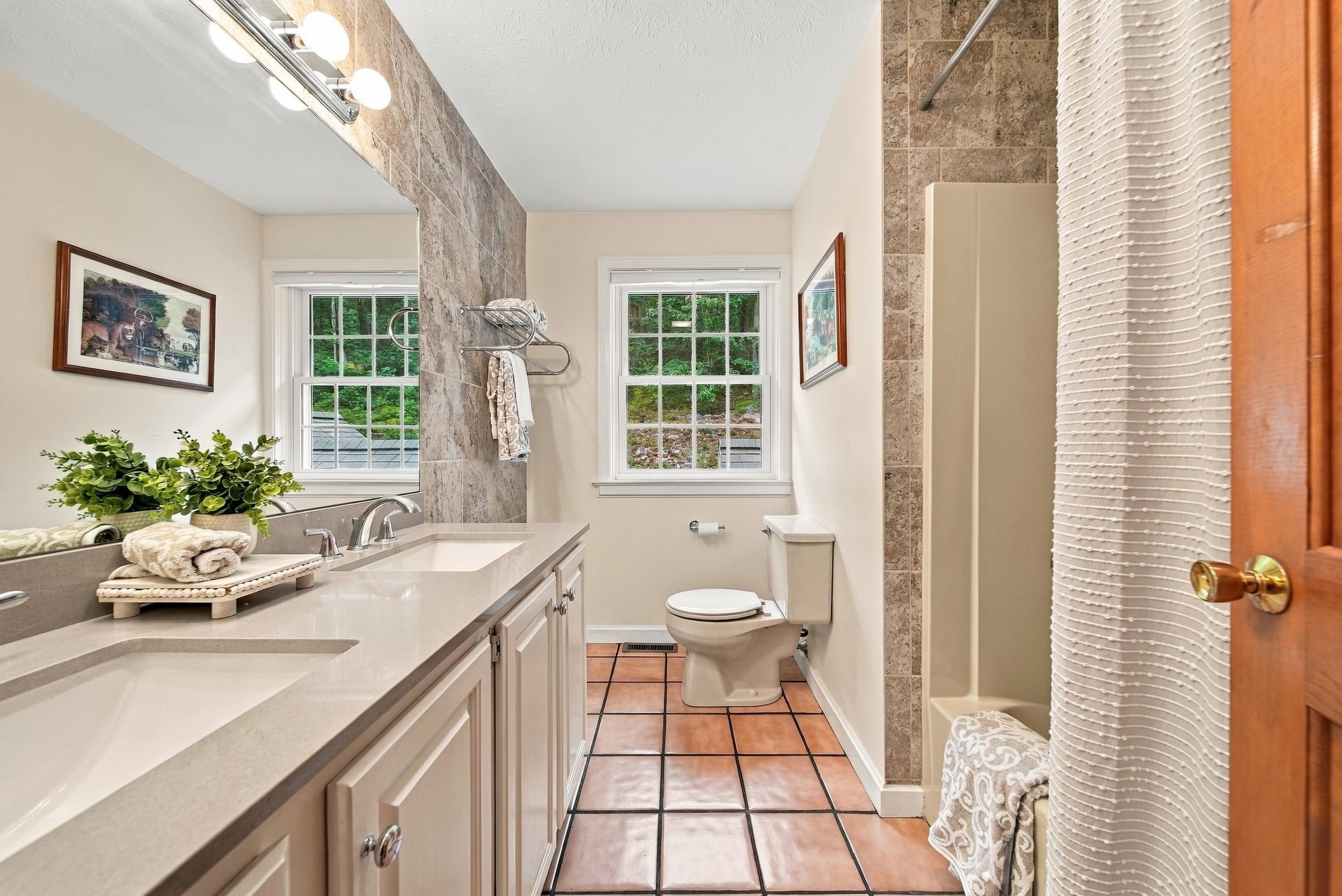Bathroom with a double-sink vanity, tiled shower, terracotta floor, and two windows overlooking greenery.