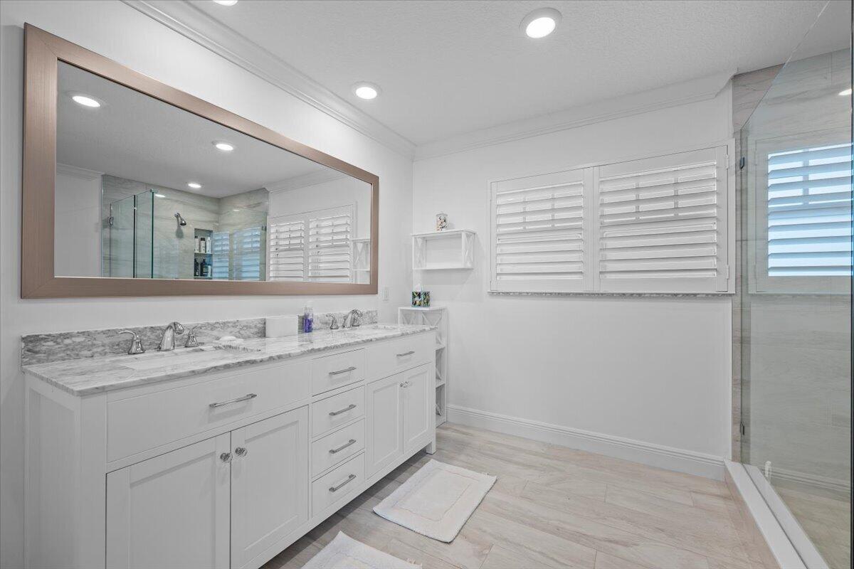 Bathroom with a marble-topped vanity, oversized mirror, plantation shutters, and glass-enclosed shower.