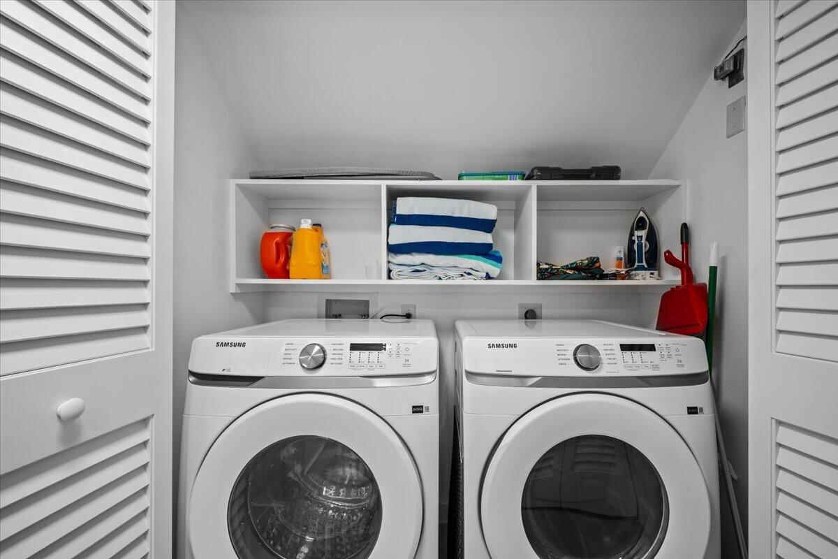 Laundry room with matching Samsung washer and dryer beneath shelves of folded towels and supplies.