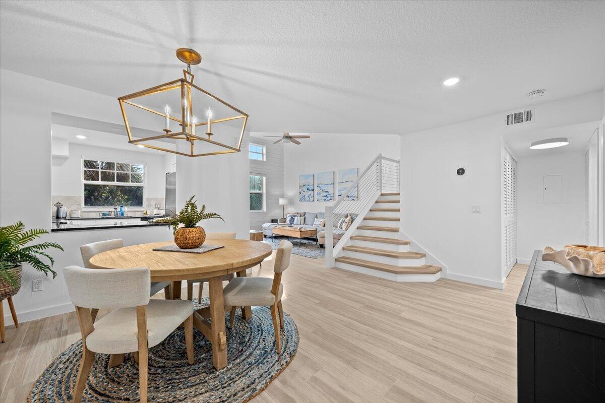 Open-plan dining area with a round table, brass pendant chandelier, kitchen, living room, and staircase.