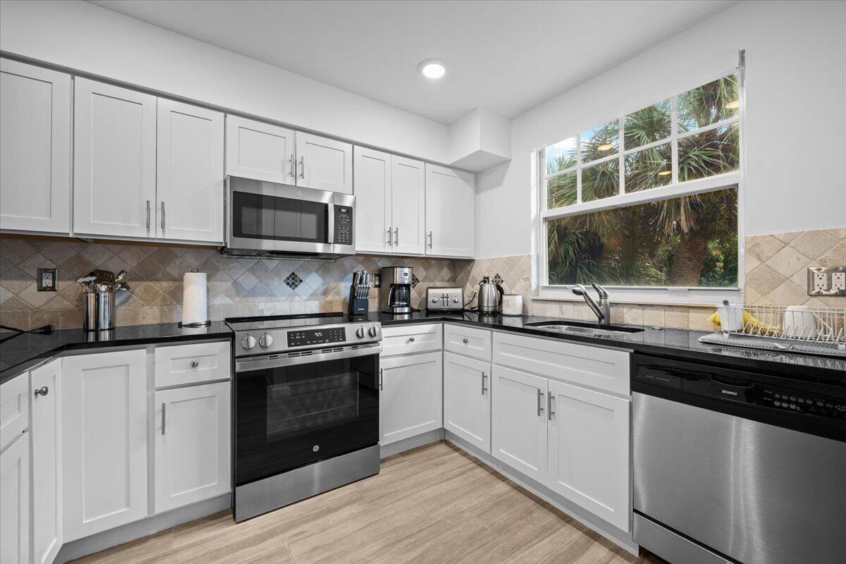 Kitchen with white cabinets, stainless steel appliances, black countertops, and a large window overlooking palm trees.