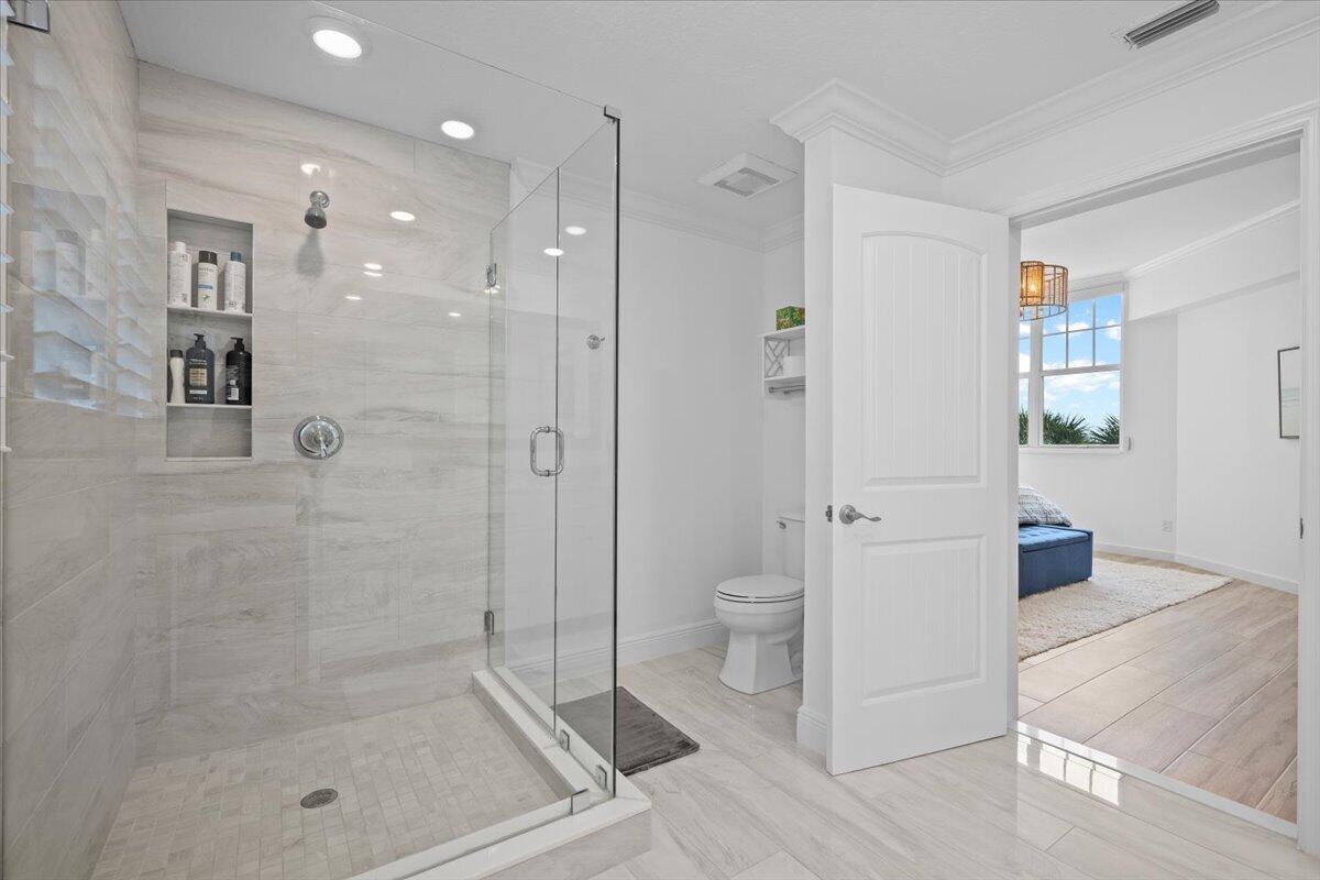 Modern bathroom with a glass walk-in shower and pale tile, opening onto a bedroom.