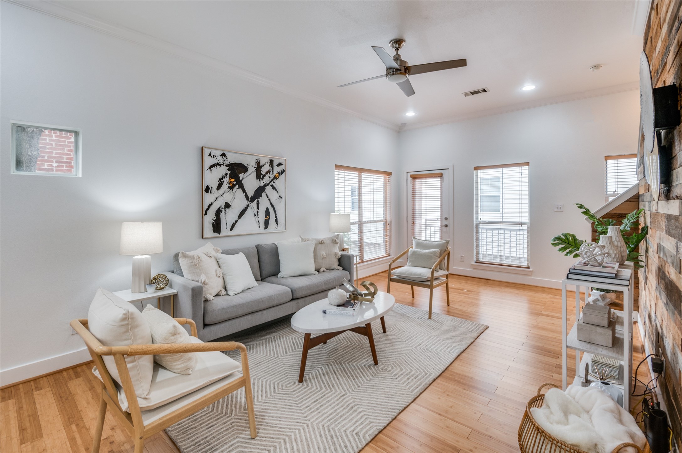 Staged living room with a gray sofa, wood floors, large windows, and a wood accent wall.