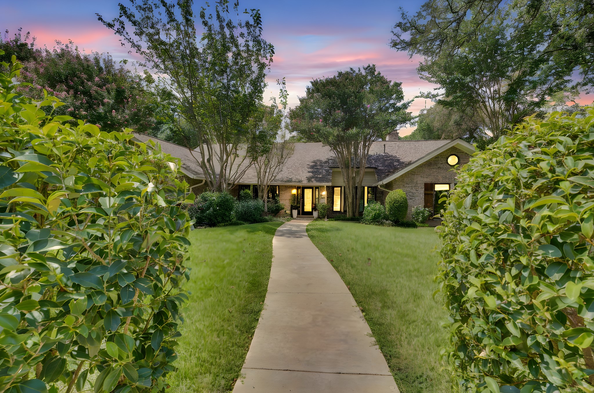 Brick home framed by leafy shrubs, with a curving walkway and warmly lit entrance at dusk.