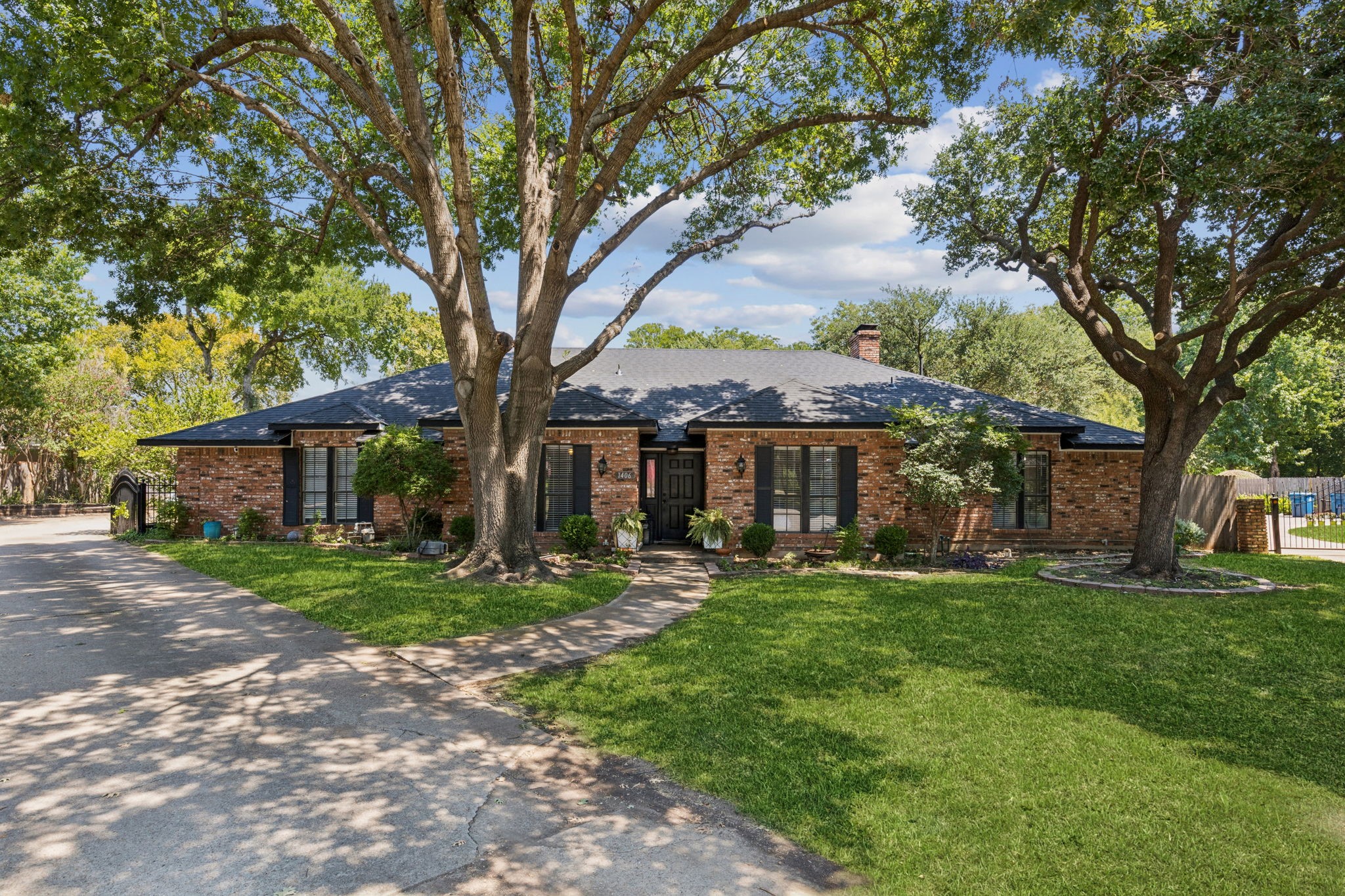 Single-story brick house framed by mature trees, with a broad lawn and curved front walkway.