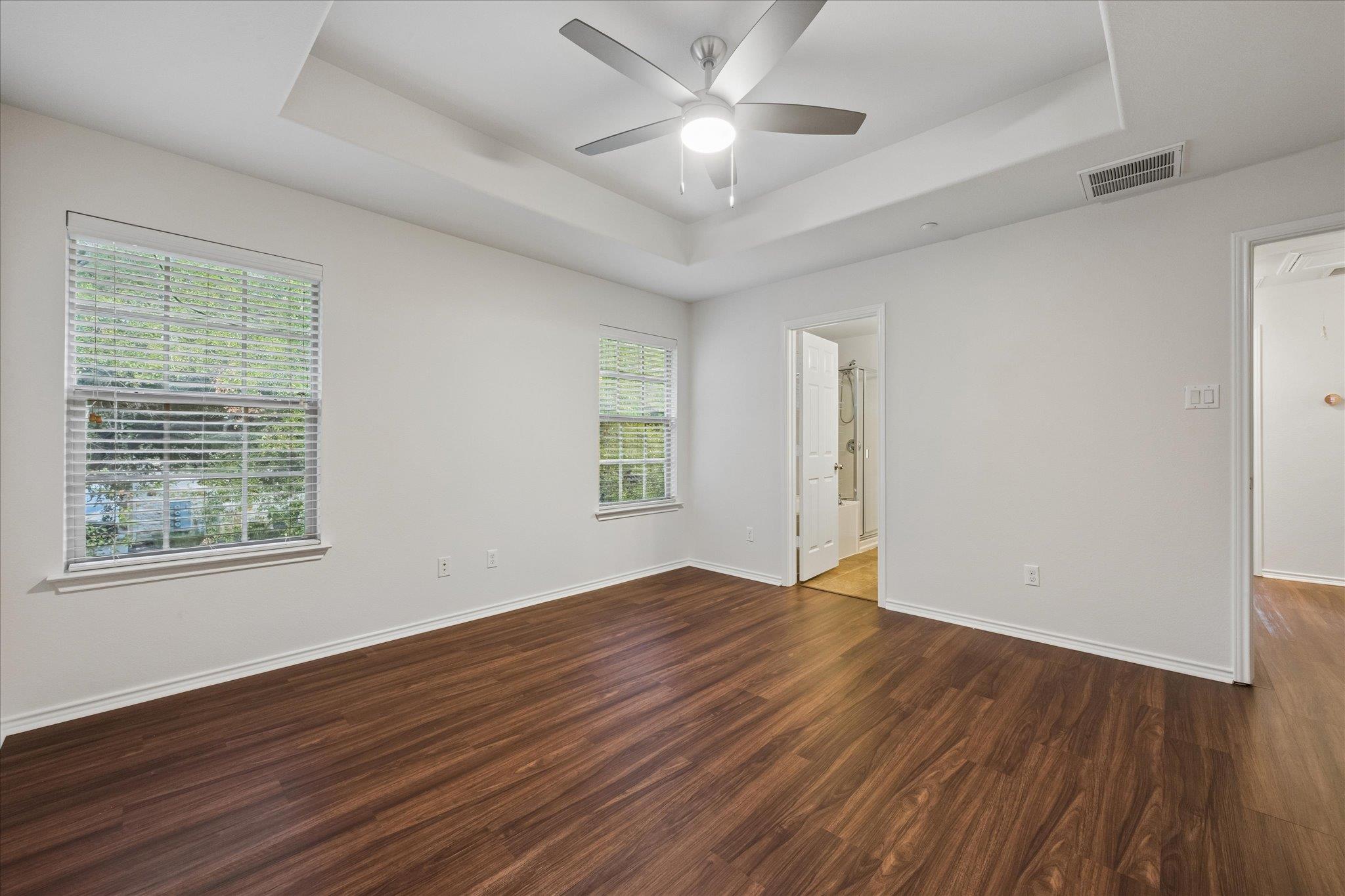 Empty bedroom with dark wood floors, two shuttered windows, and a tray ceiling fan.