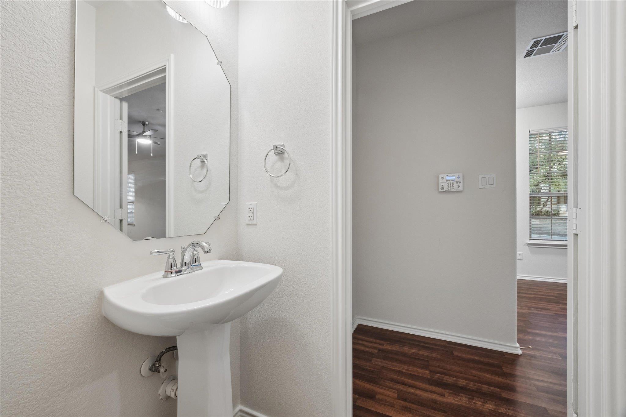 Bathroom with a white pedestal sink and angular mirror opening onto a dark wood-floored hall.