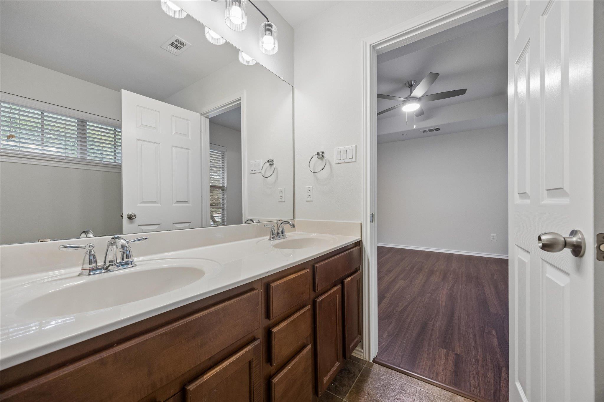Bathroom with a double-sink vanity and dark wood cabinets, opening to a room with a ceiling fan.