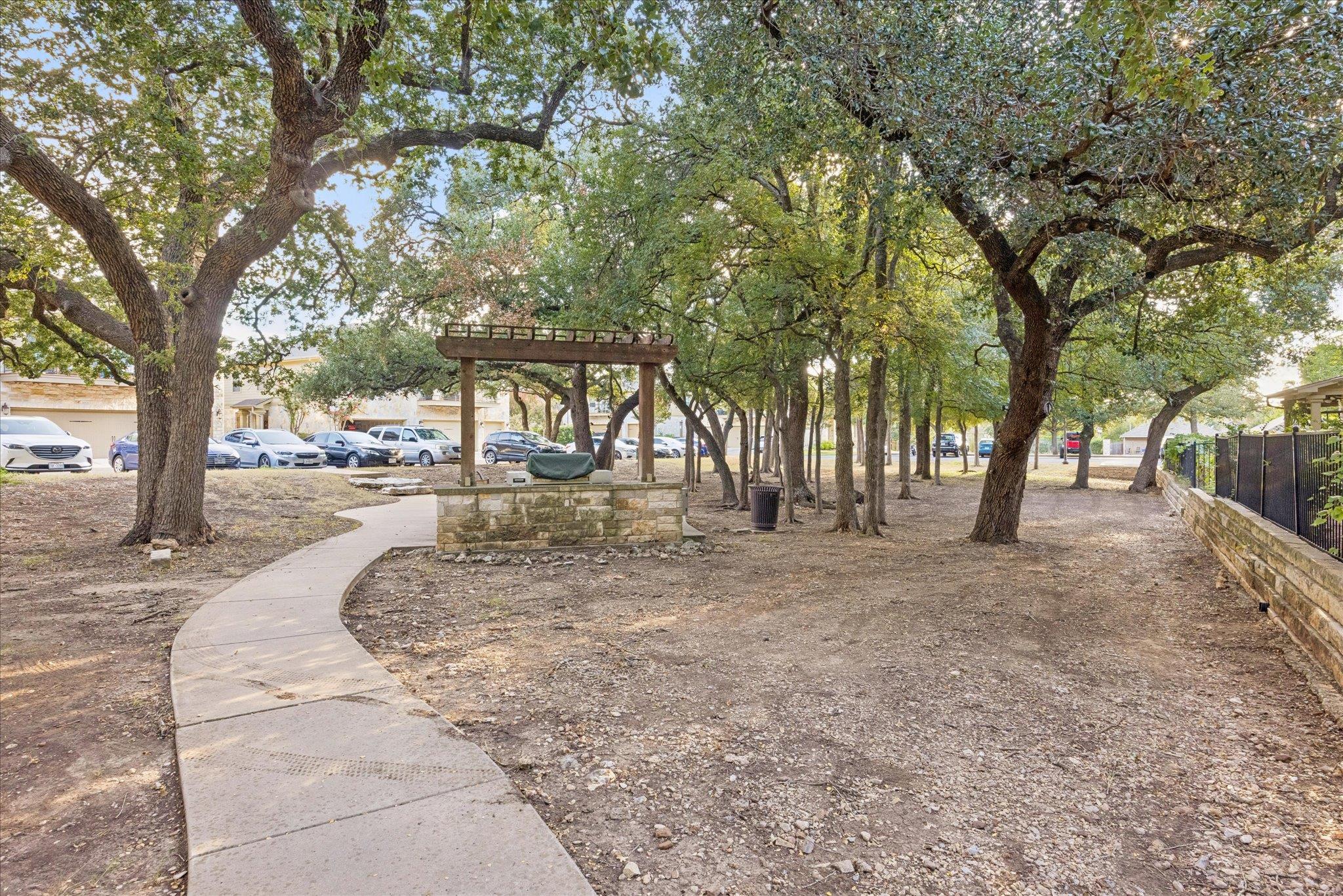 Shaded courtyard with mature oak trees, a curved walkway, and a stone seating area beneath a pergola.