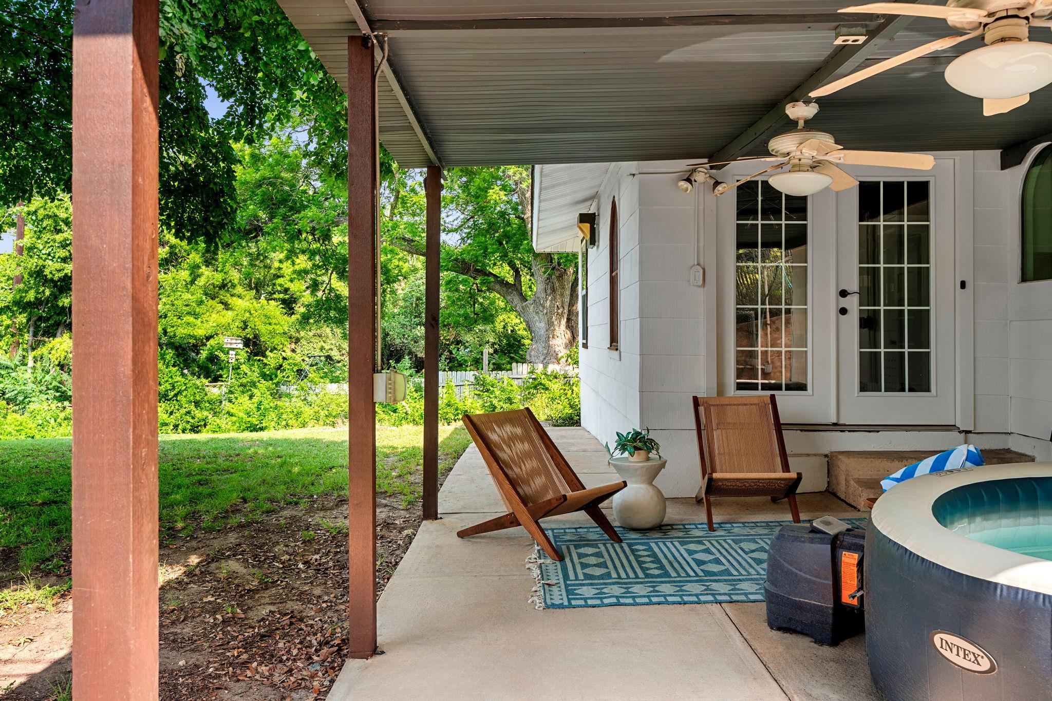 Covered backyard patio with wooden lounge chairs, ceiling fans, and an inflatable hot tub beside a leafy yard.