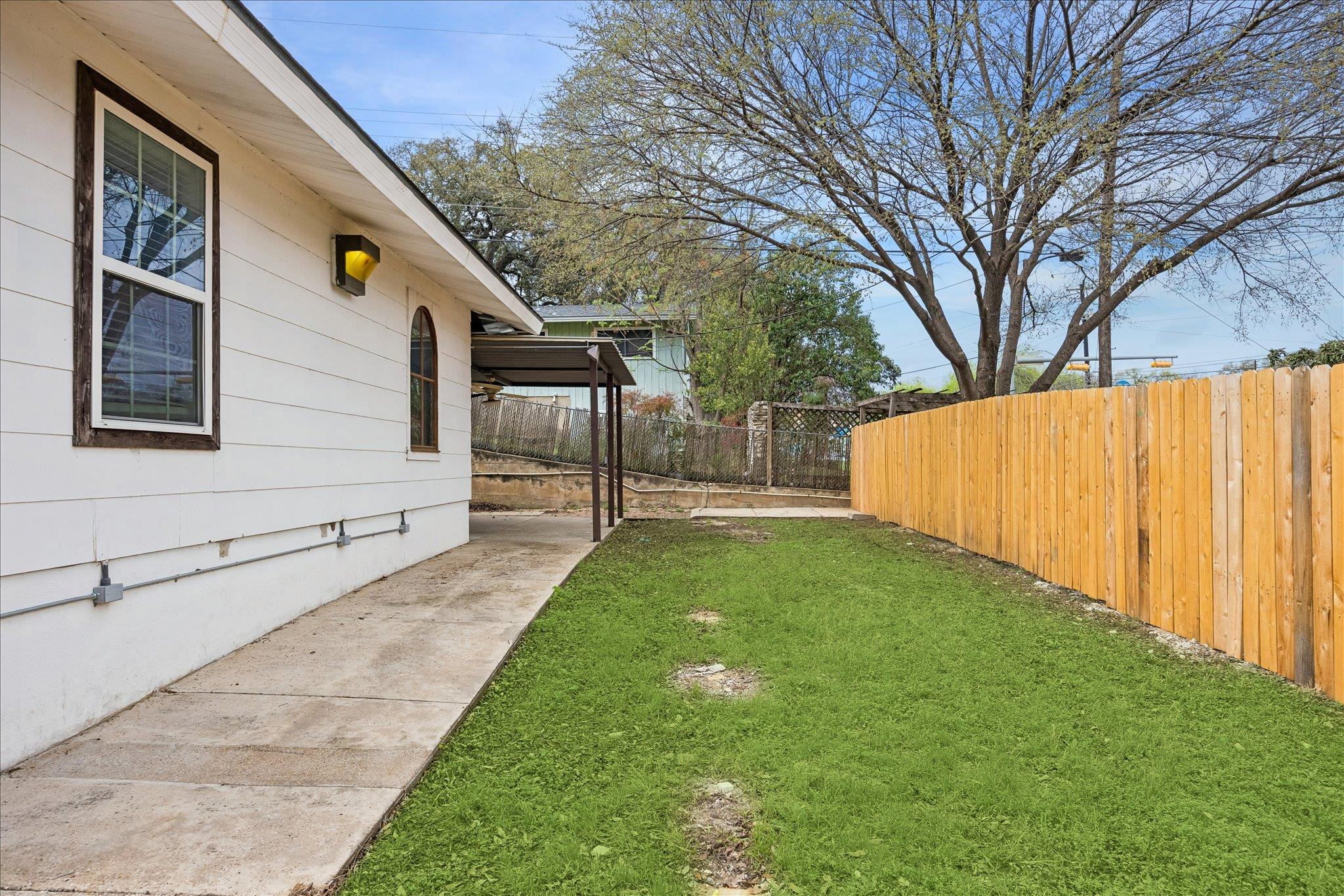 Side yard with a green lawn, concrete walkway, covered patio, and tall wooden fence.
