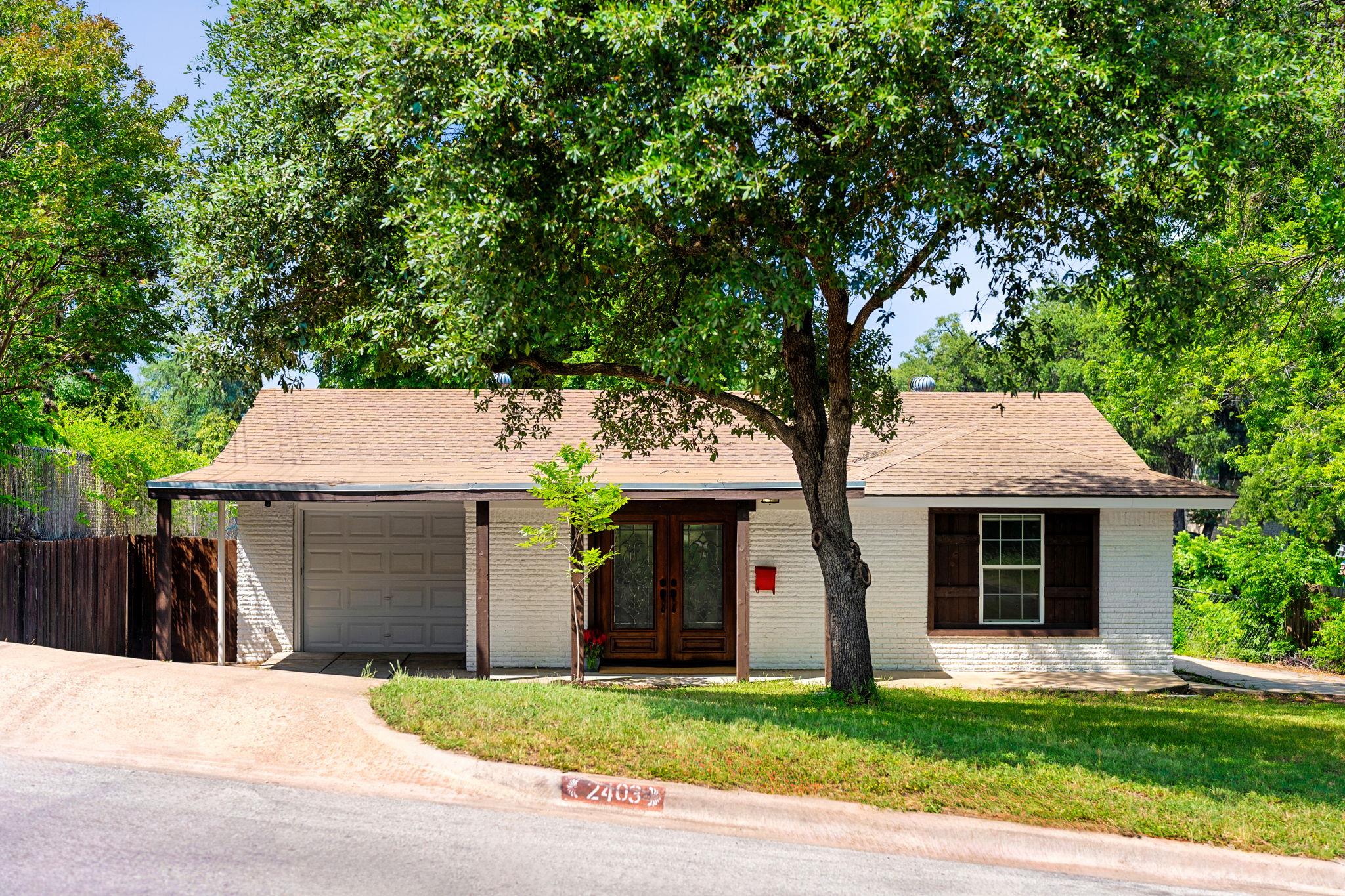 Single-story white brick house with a garage and dark wood entry beneath a large shade tree.