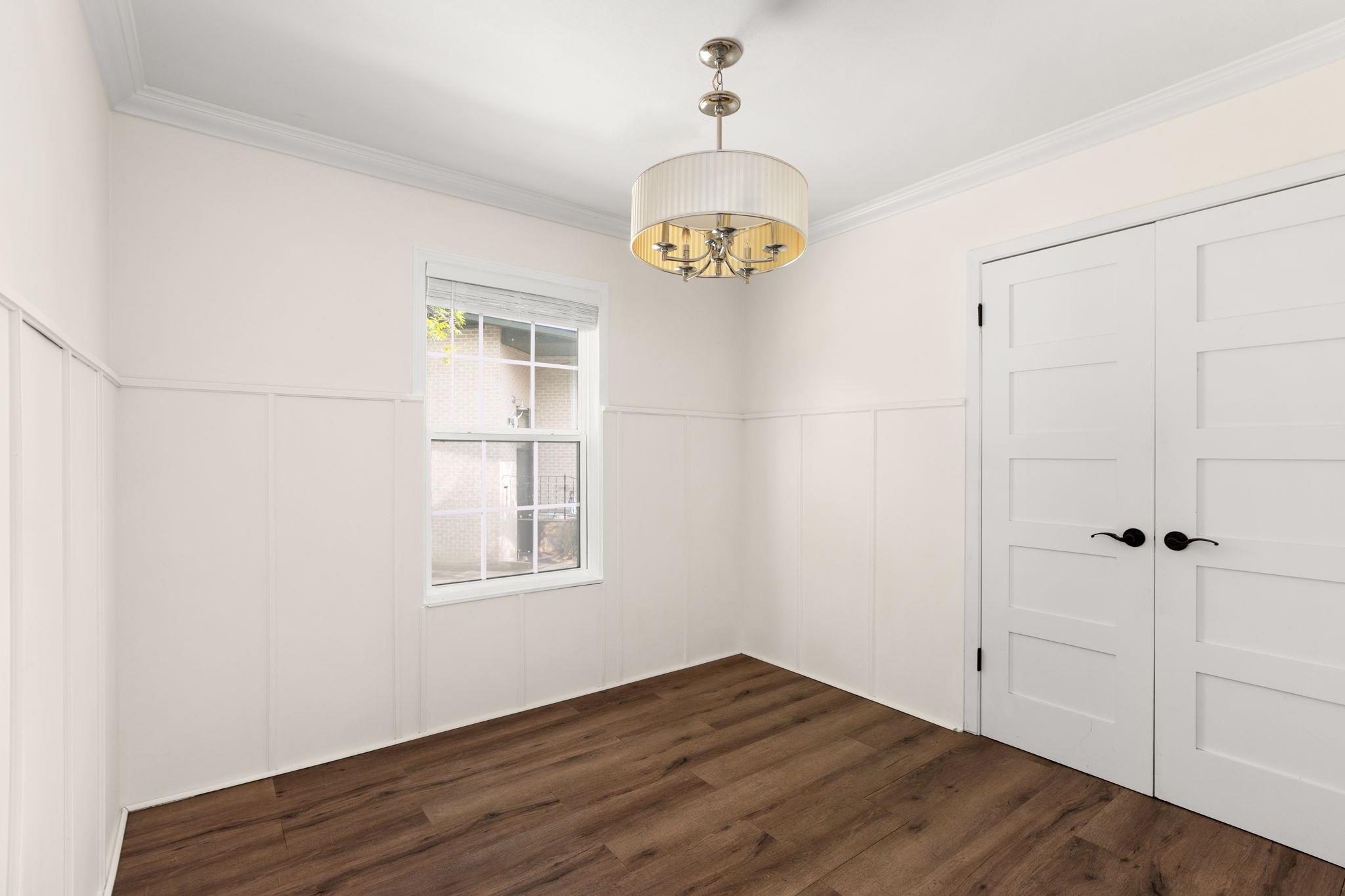 Empty room with white paneled walls, dark wood flooring, a window, double doors, and a brass chandelier.