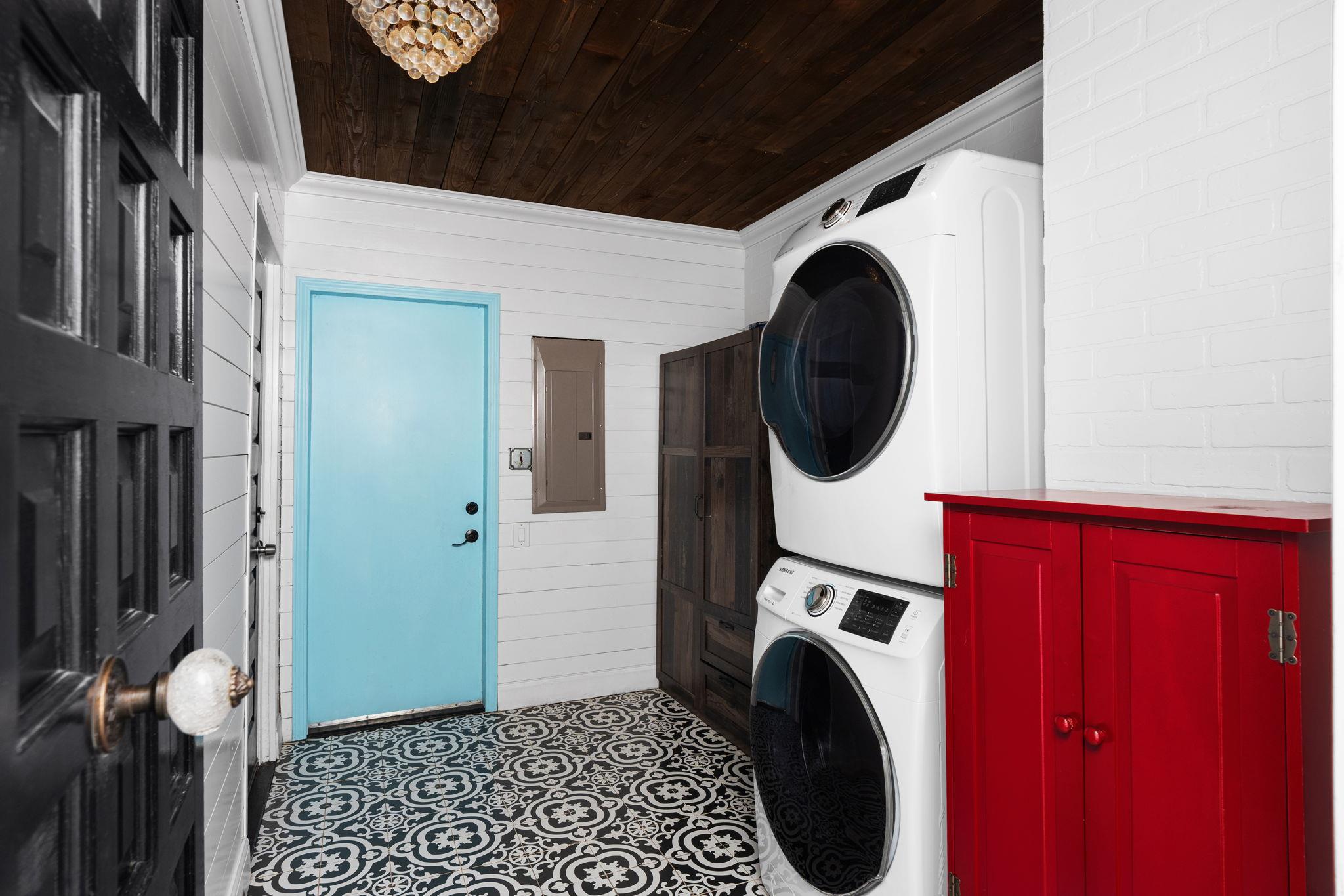 Laundry room with stacked washer and dryer, a bright blue door, red cabinet, and patterned tile floor.
