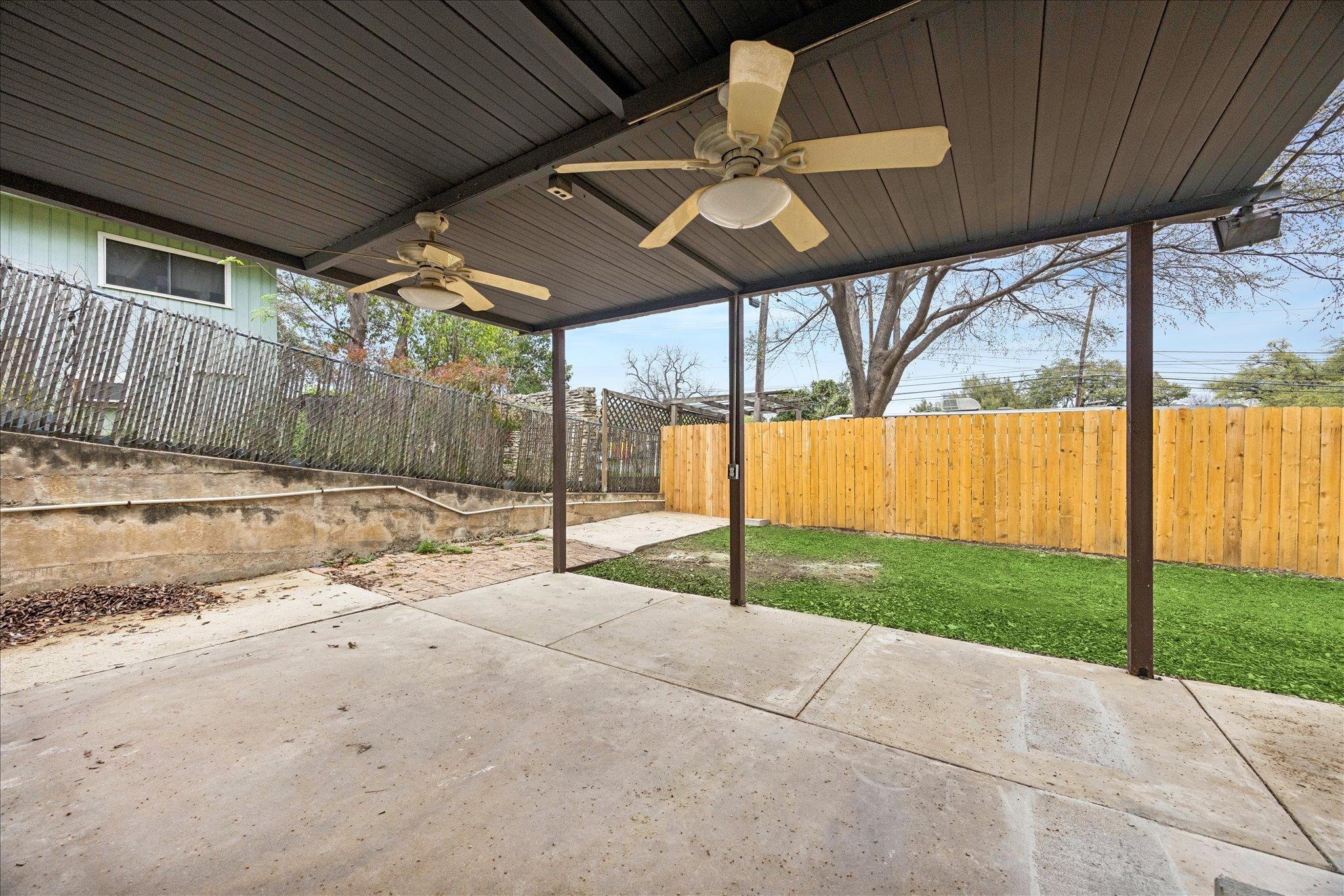 Covered concrete patio with two ceiling fans overlooking a fenced grassy backyard.