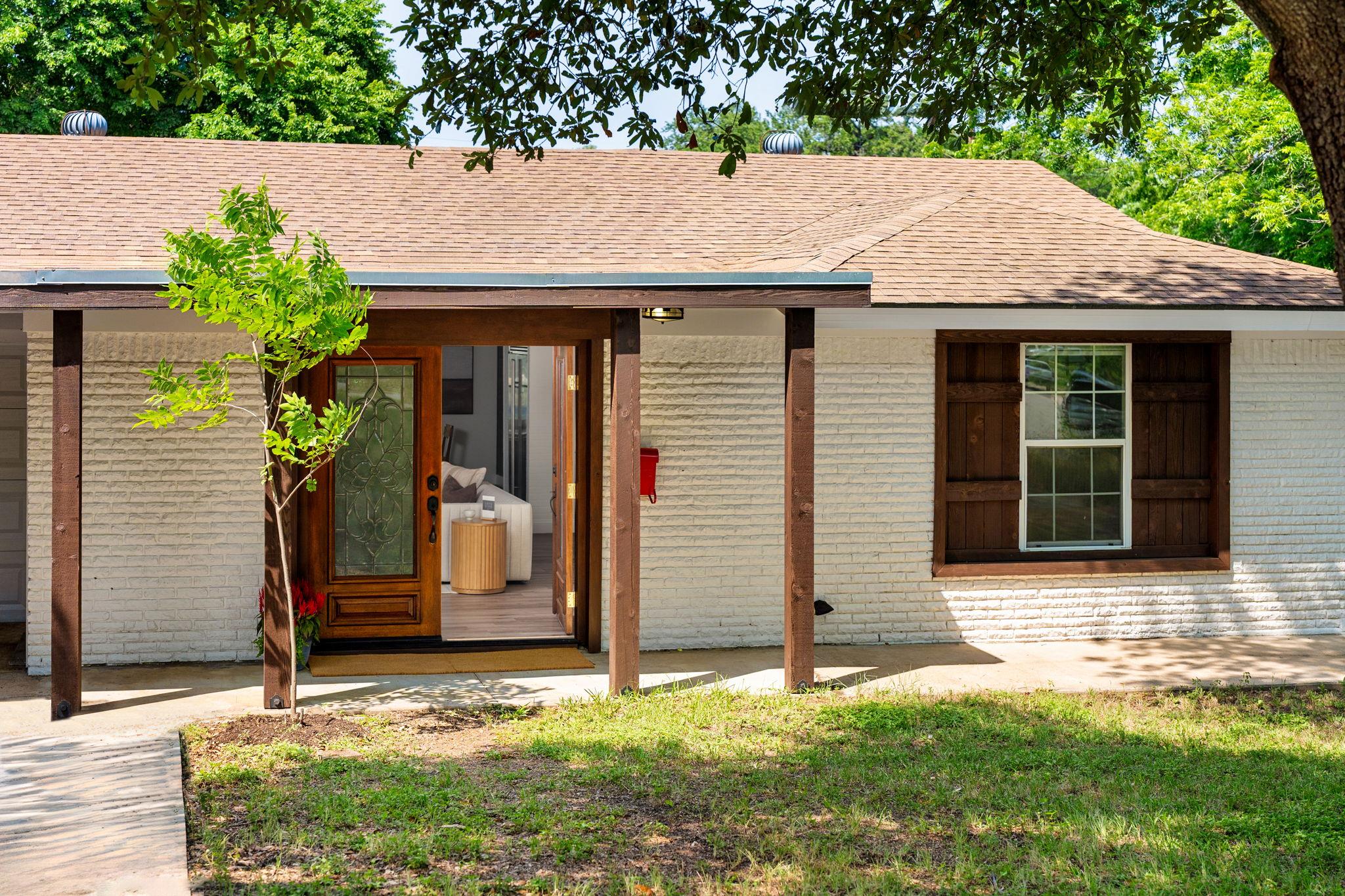 Single-story brick home with a covered entry, open wooden front door, and mature trees in the yard.