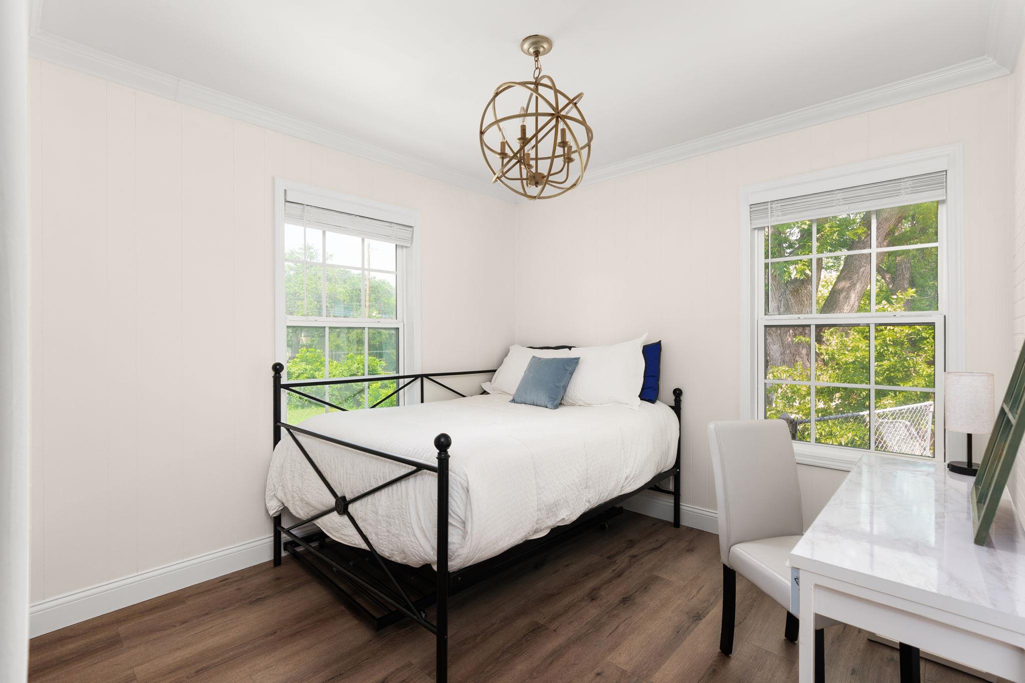 Bedroom with a black metal bed, leafy views through two windows, and a brass globe chandelier.