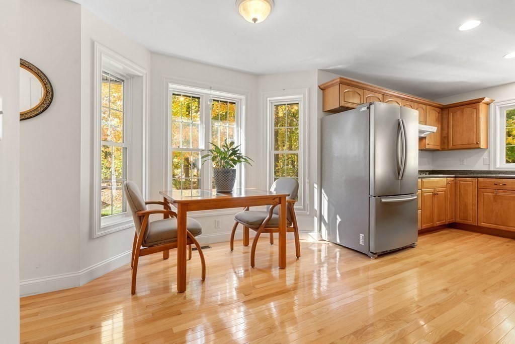 Kitchen dining area with wood cabinets, stainless-steel refrigerator, hardwood floors, and tall windows overlooking autumn foliage.