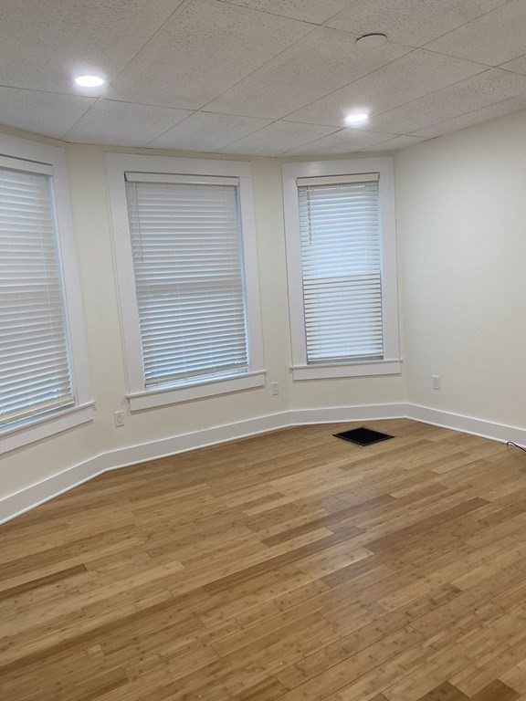 Vacant room with light wood floors and three windows with blinds.