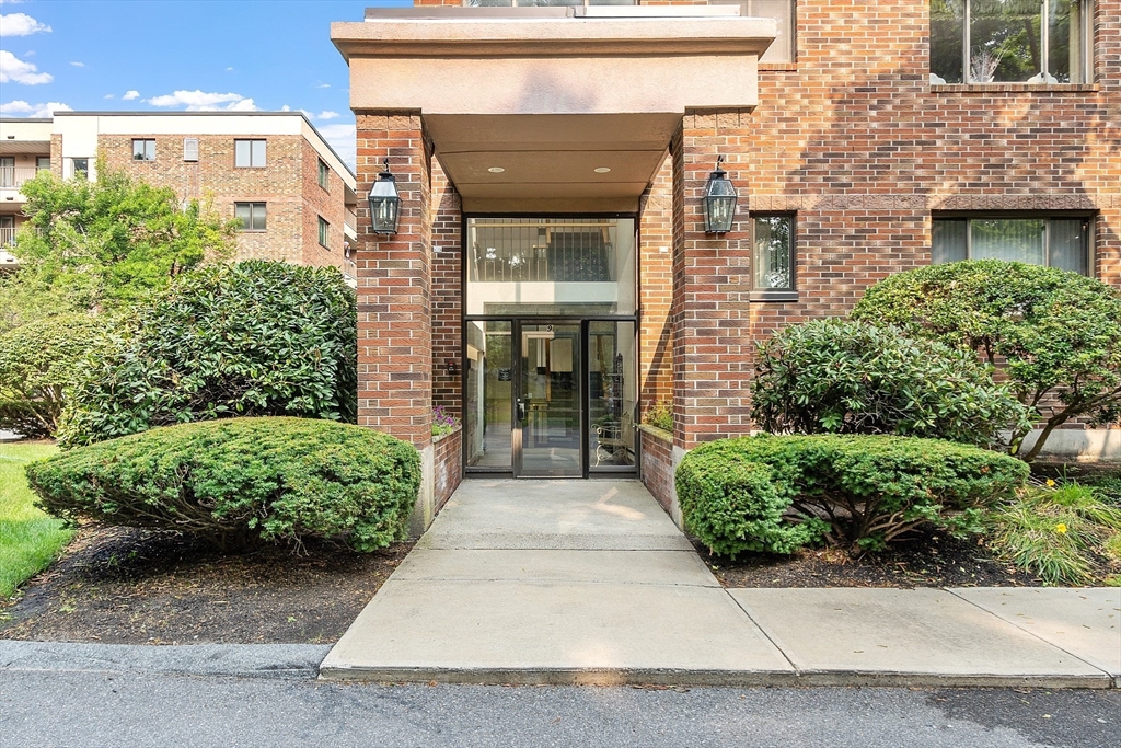 Brick residential building entrance with a glass door, covered portico, and landscaped shrubs.