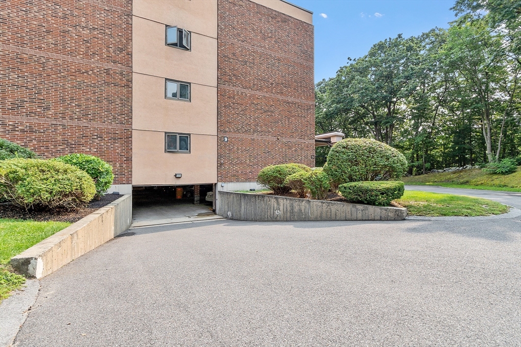 Brick condominium exterior with a covered parking entrance, shrubs, and a paved driveway.