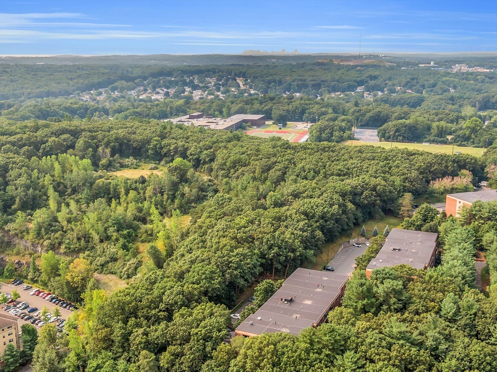 Aerial view of wooded surroundings, nearby buildings, and a sports field and track in the distance.