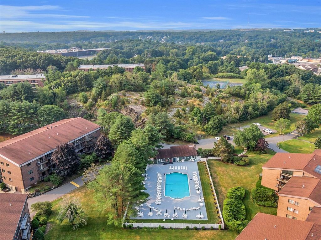Aerial view of a condominium complex with a central pool, landscaped grounds, and wooded hills beyond.