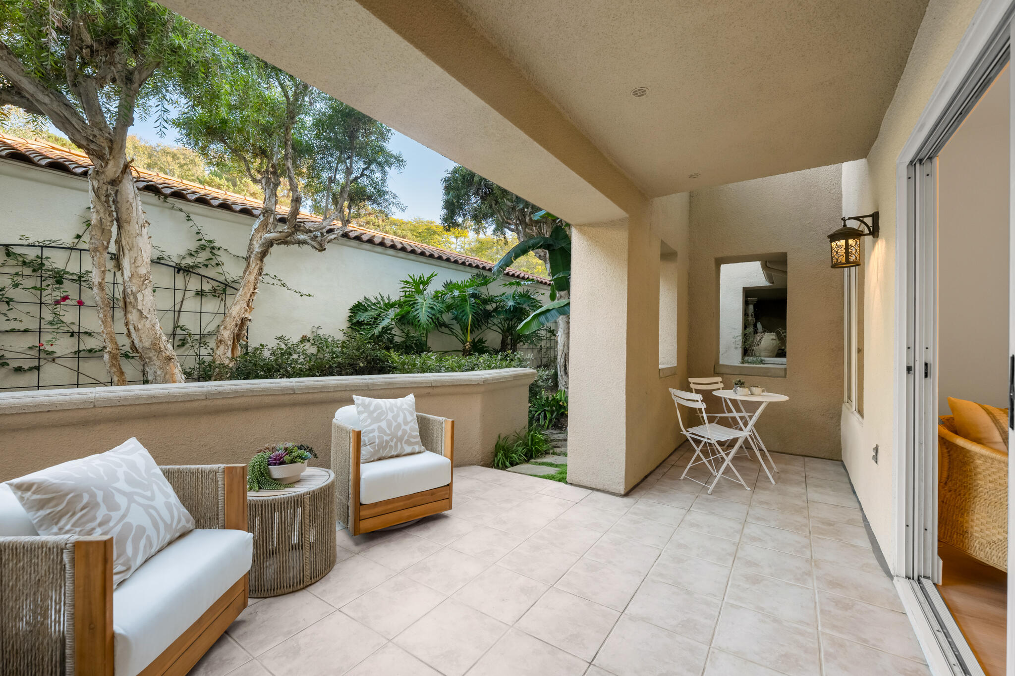 Covered patio with wicker seating and a café table beside a leafy courtyard garden.