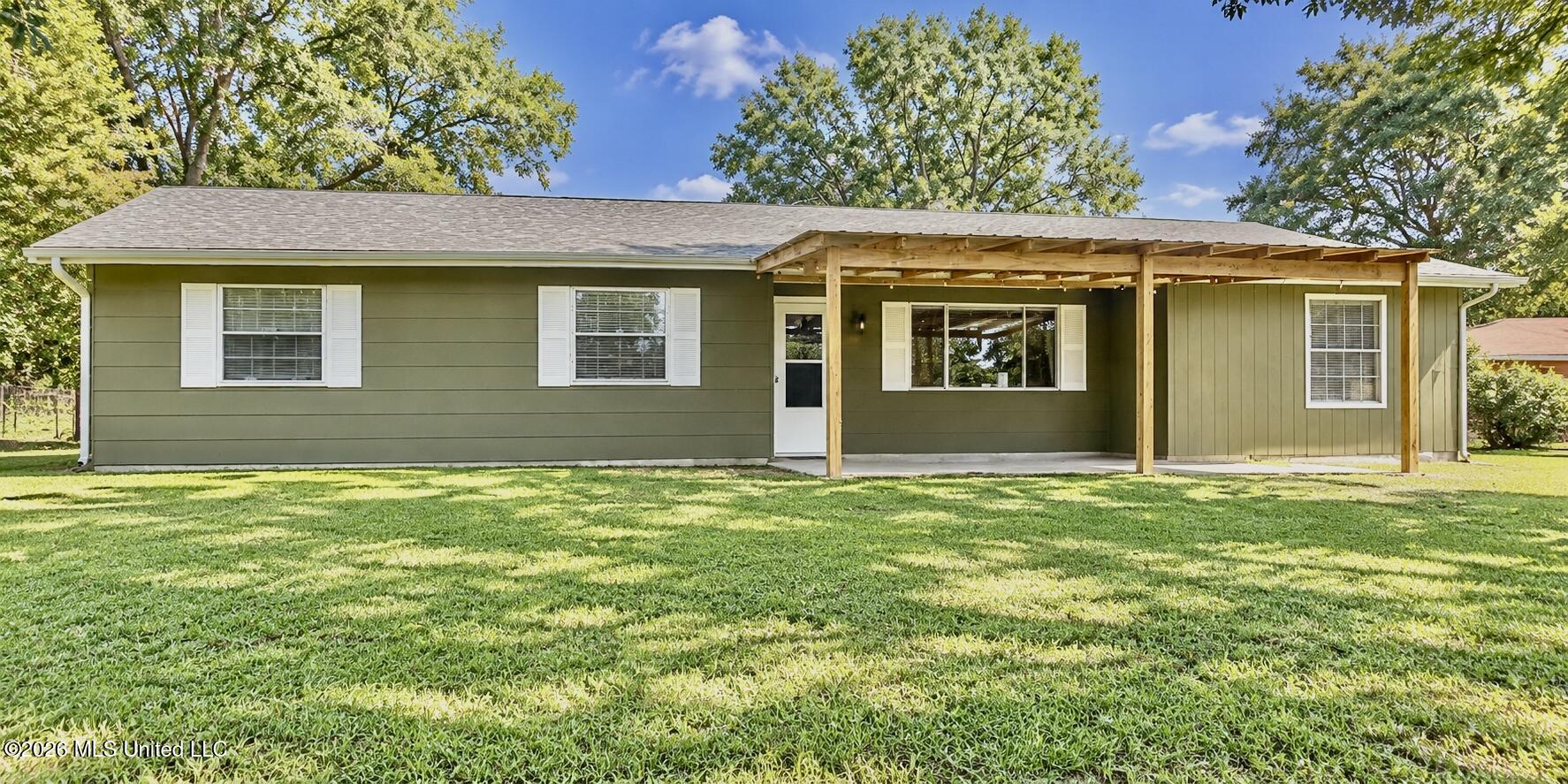 Single-story green home with white shutters and a timber-covered patio overlooking a broad lawn.