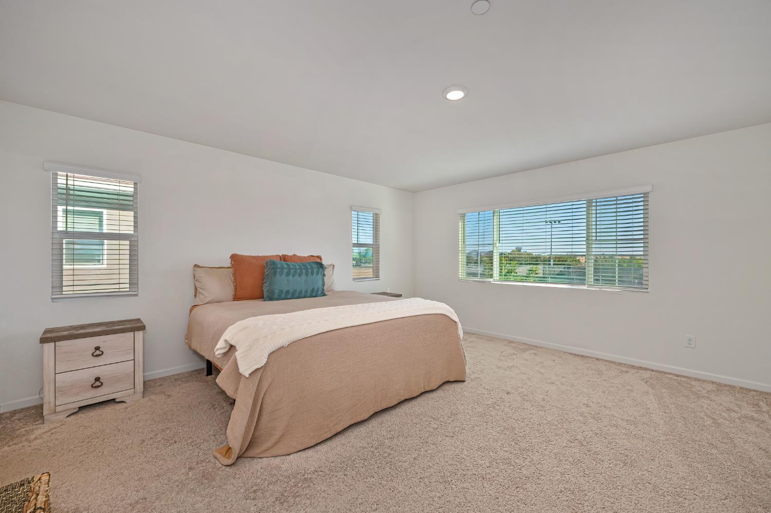 Furnished bedroom with a neutral bed, colorful accent pillows, beige carpet, and wide windows with blinds.