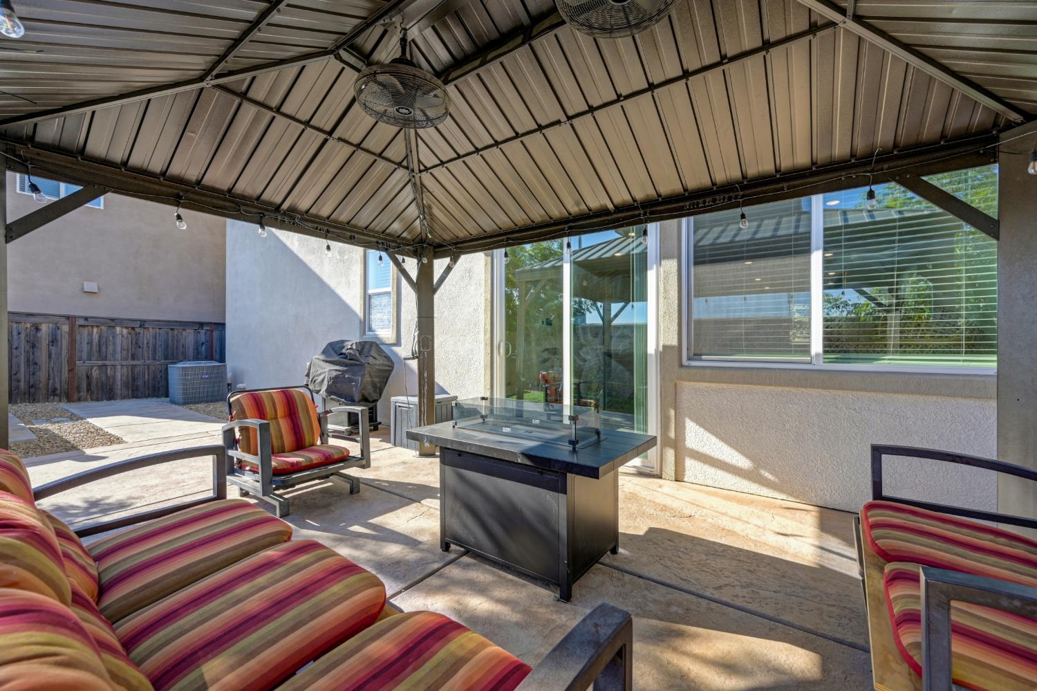 Covered patio with striped lounge seating and a central fire table beneath a metal pavilion.