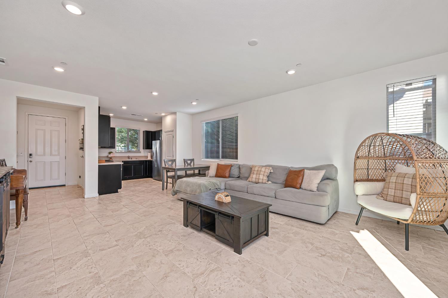 Open-plan living room with gray sofa, wicker chair, tiled floors, and dark-cabinet kitchen.