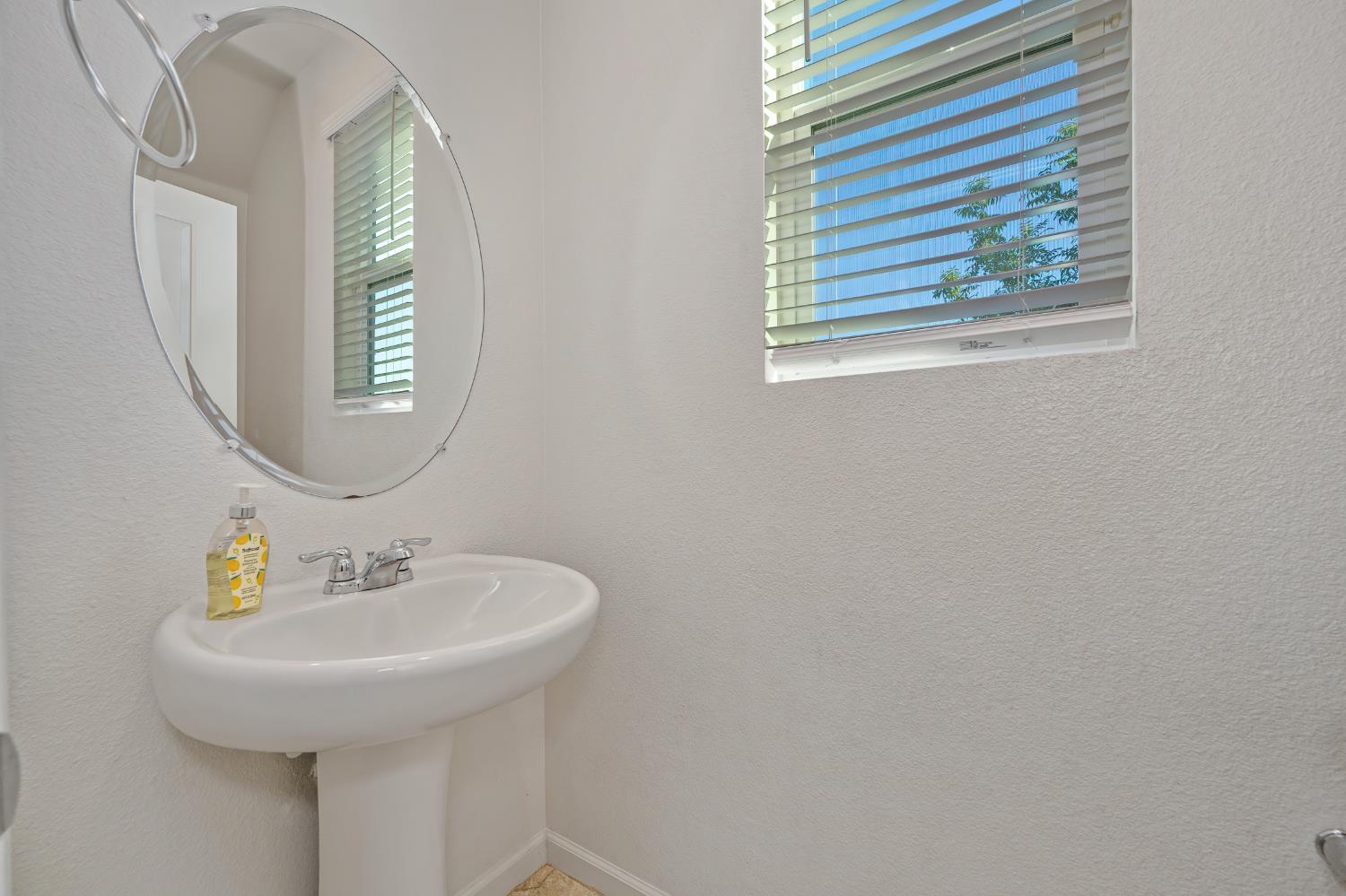 Bathroom with a pedestal sink, large oval mirror, and window with blinds.