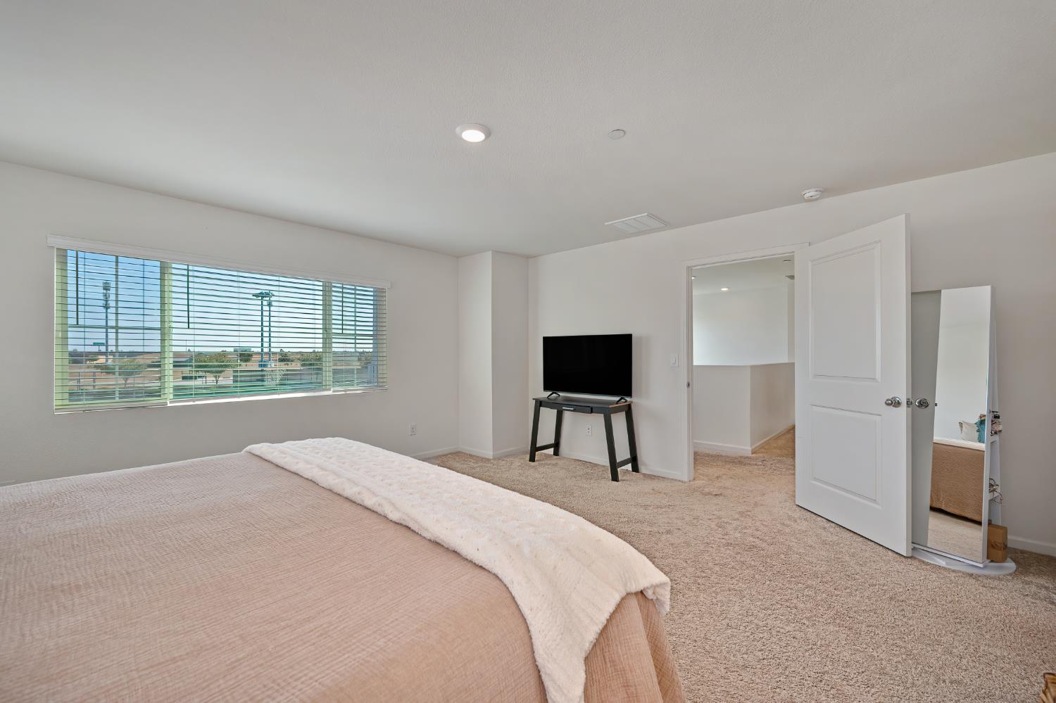 Bedroom with a large bed, wide window with blinds, TV, and full-length mirror.