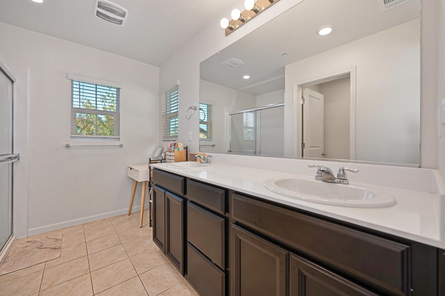 Bright bathroom with a double-sink vanity, dark cabinets, large mirror, and glass-enclosed shower.