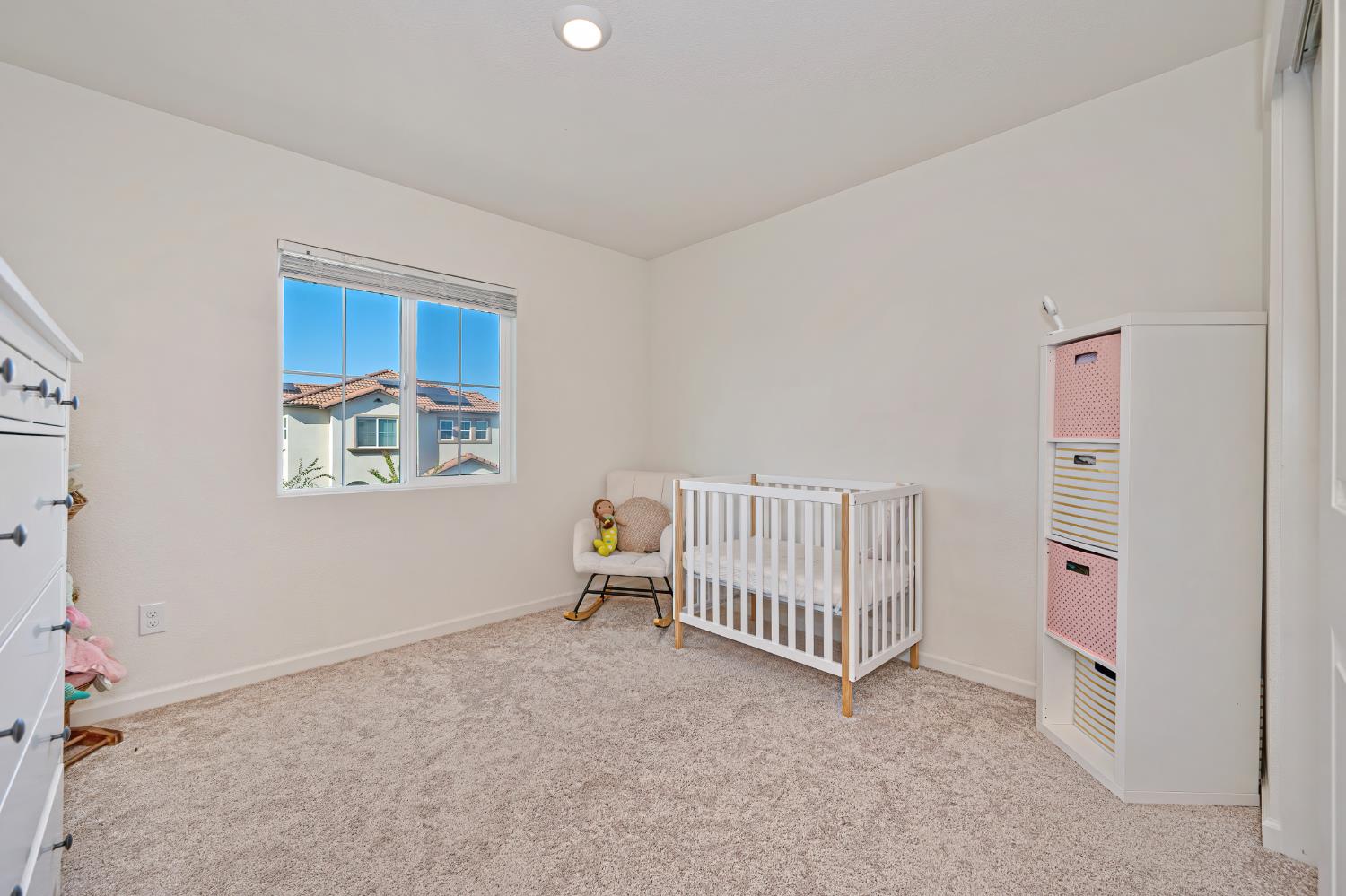 Nursery bedroom with a white crib, rocking chair, and colorful storage bins beside a sunny window.
